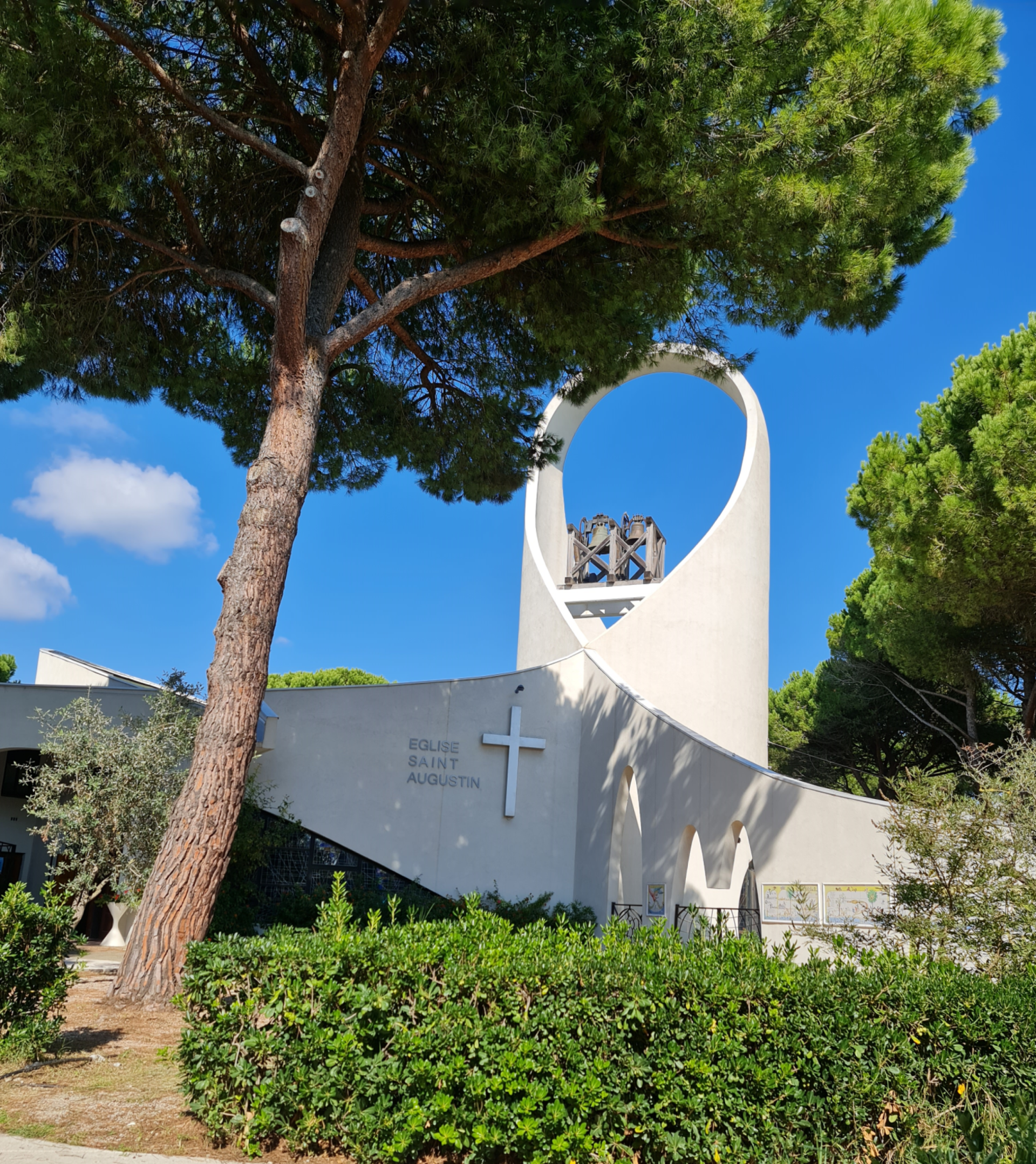 weißer Glockenturm und Gebäude einer Kirche, moderne Archtektur, an der Mauer steht der Name der Kirche Sankt Augustinus, Turm in Form 2 er über dem Kopf verschränkter Arme, davor grüne Büsche und eine Pinie