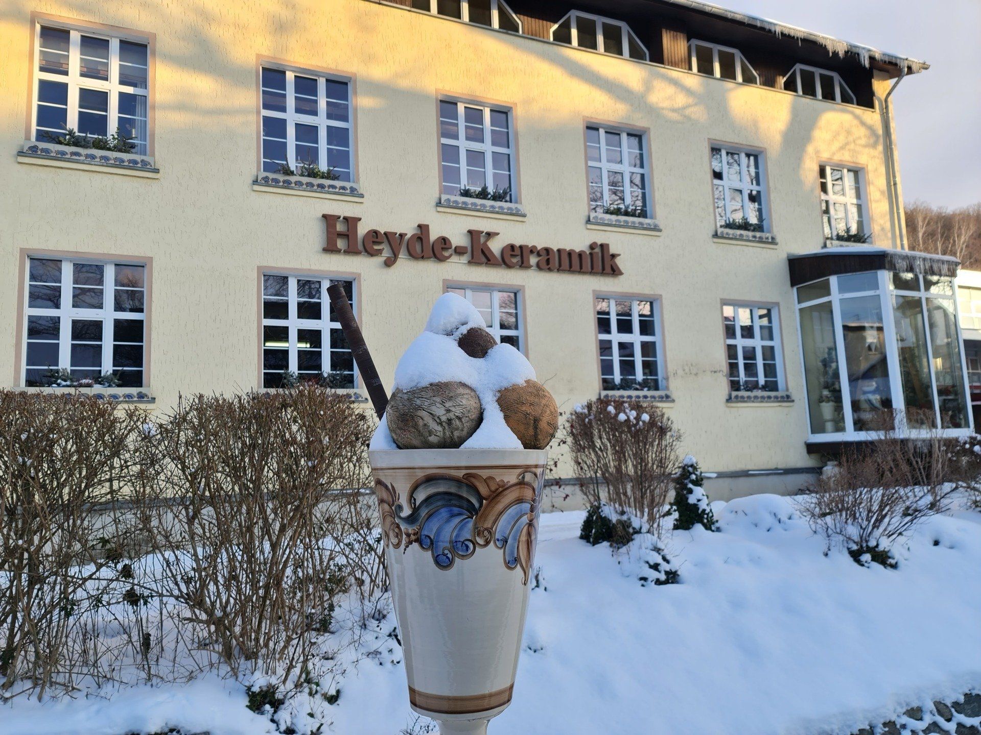 Heyde Keramik in Jahnsdorf - schon vor der Tür der Fabrikation sieht man die tollsten Exemplare der hiesigen Töpferkunst Haus in hellgelb, viele Fenster, ein ehemaliges Fabrikgebäude mit Buchstaben Heyde Keramik vorn dran. Im Vordergrund steht ein riesiger Eisbecher aus Keramik in Natur mit Muster aus blau und braun, Eiskugeln und Strohhalm sind verschiedenfarbig braun wie Schokolade
