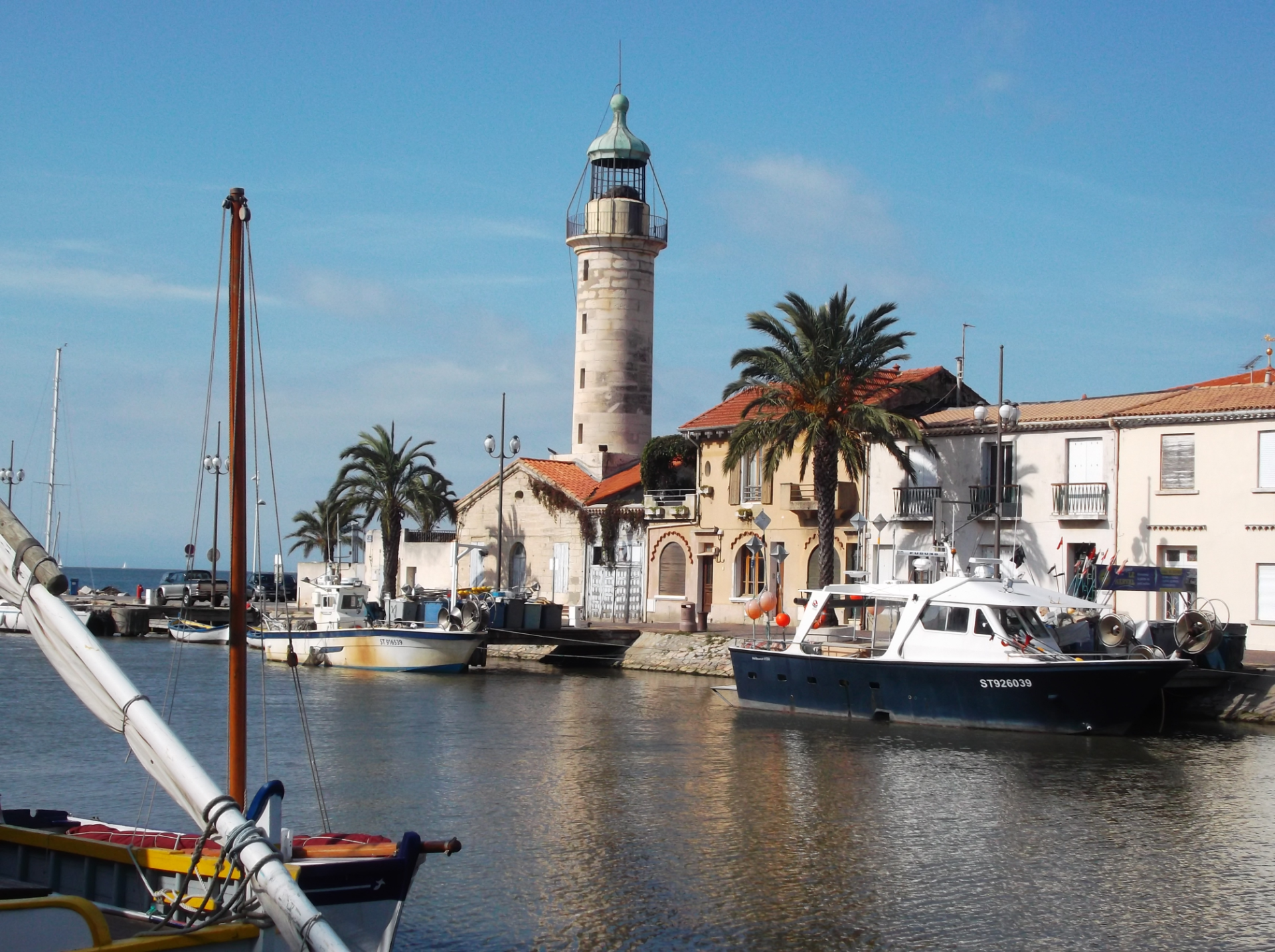Leuchtturm und Häuser mit Palmen davor auf der rechten Seite, mittig der Kanal mit Booten auf der rechten Seite unter blauem Himmel