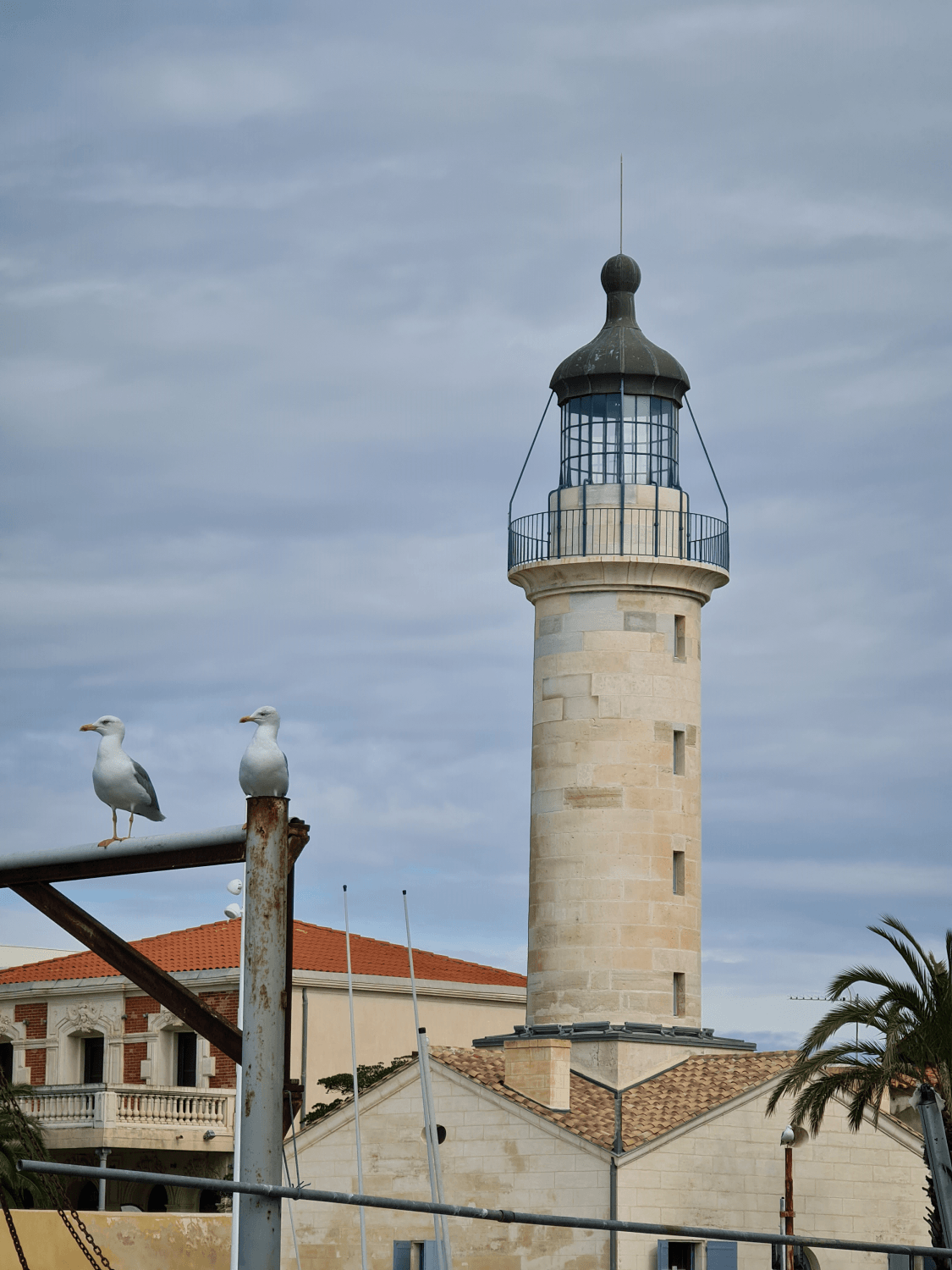 2 Möwen sitzen auf einer Stange vor dem Leuchtturm von Le Grau du Roi, beide Steine, blau-grauer Himmel, im Hintergrund die Tourist-Information