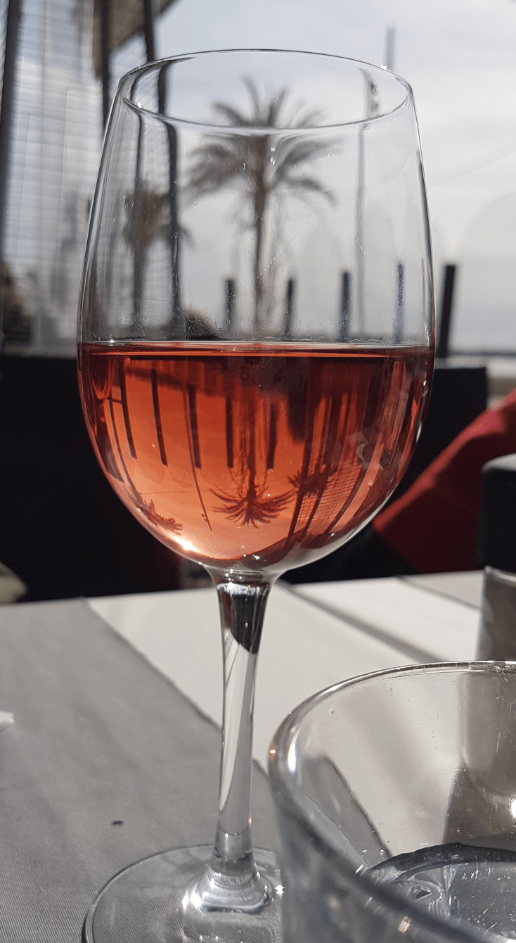Weinglas halb gefüllt mit rosé Wein steht im #'Restaurant in Le Grau du Roi auf dem Tisch im Glas in der oberen Glashälfte ist eine Palme zu sehen die sich im Wein spiegelt