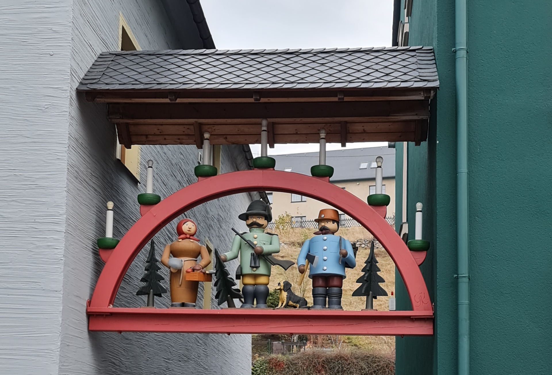 Tradition um Weihnachten - Schwibbogen - hier zwischen 2 Häusern angebracht mit typischen Figuren. Schwibbogen aus Holz mit Lichtern zwischen 2 Häusern mit großen erzgebirgischen Figuren Jäger, Pilzsammlerin und Holzfäller