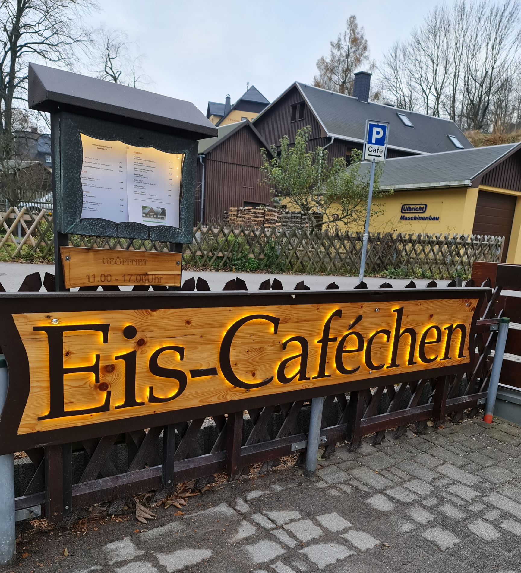 Häuschen im Hintergrund am Gartenzaun eine beleuchtete Werbung mit Eis-Caféchen Schild mit Speisekarte und Öffnungszeiten