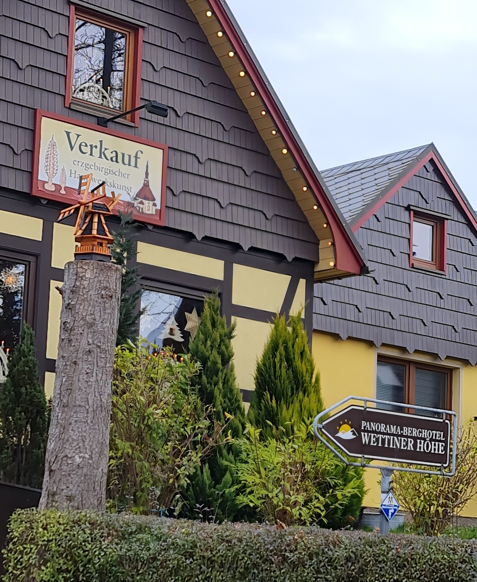 gelbes Fachwerkhaus mit Holzstamm davor oben auf dem Stamm eine Windmühle aus Holz und im Garten ein Schild zu einem Hotel