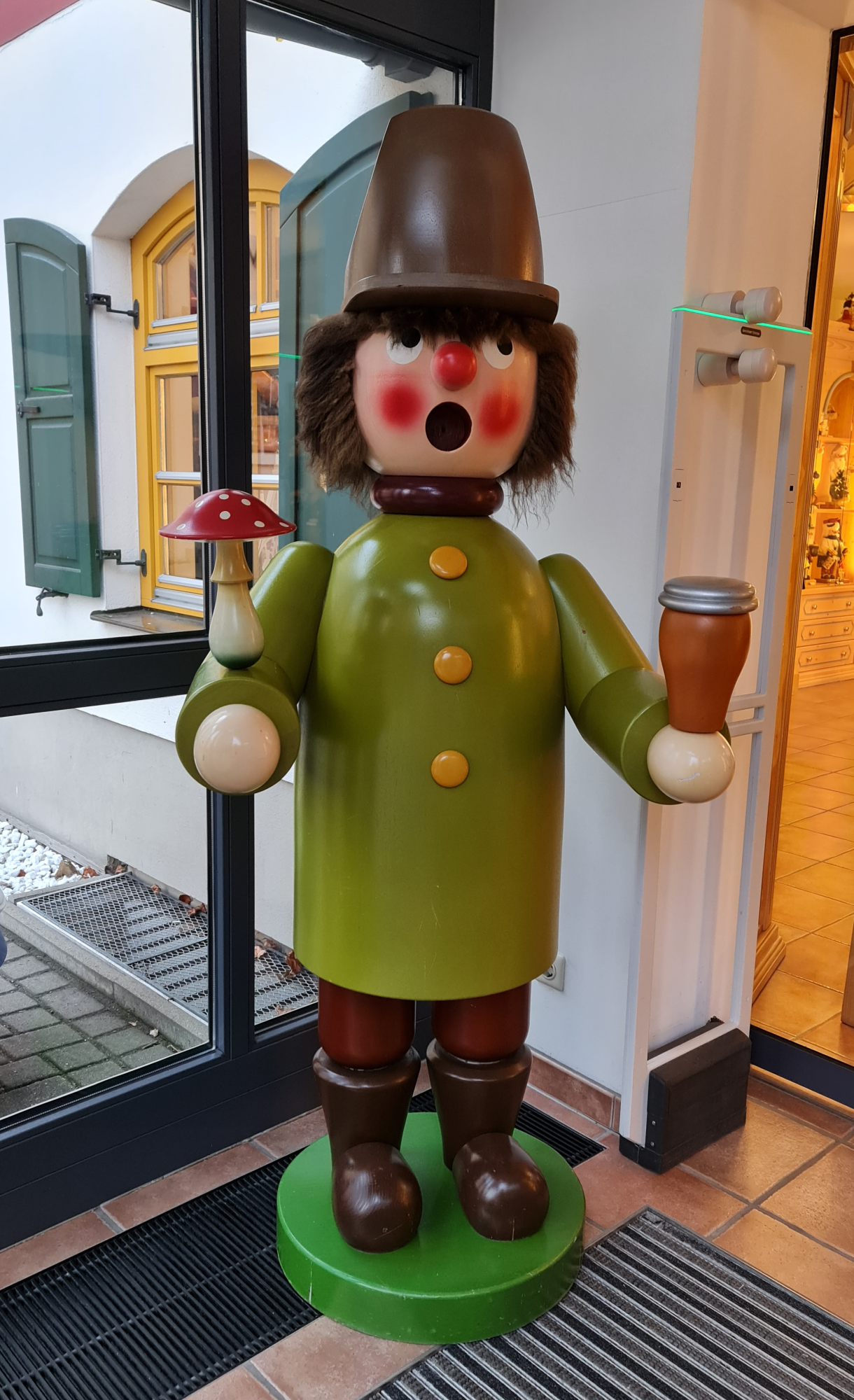 hellgrüner großer Räuchermann mit brauner Mütze und Pilz in der Hand steht in einem Vorhaus aus Glas
