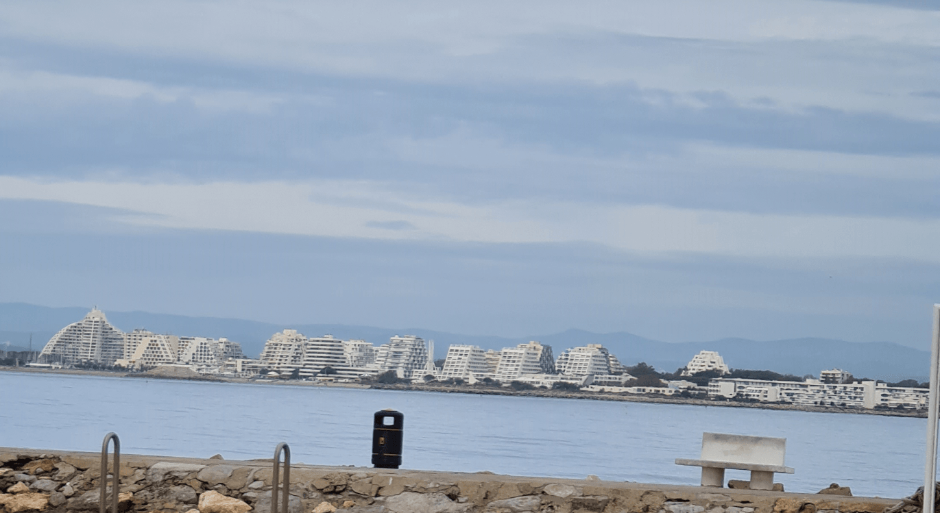im Hintergrund die Skyline einer Stadt mit weißen Gebäuden, die unterschiedliche futuristische Formen haben , davor Strand und dazwischen das Mittelmeer