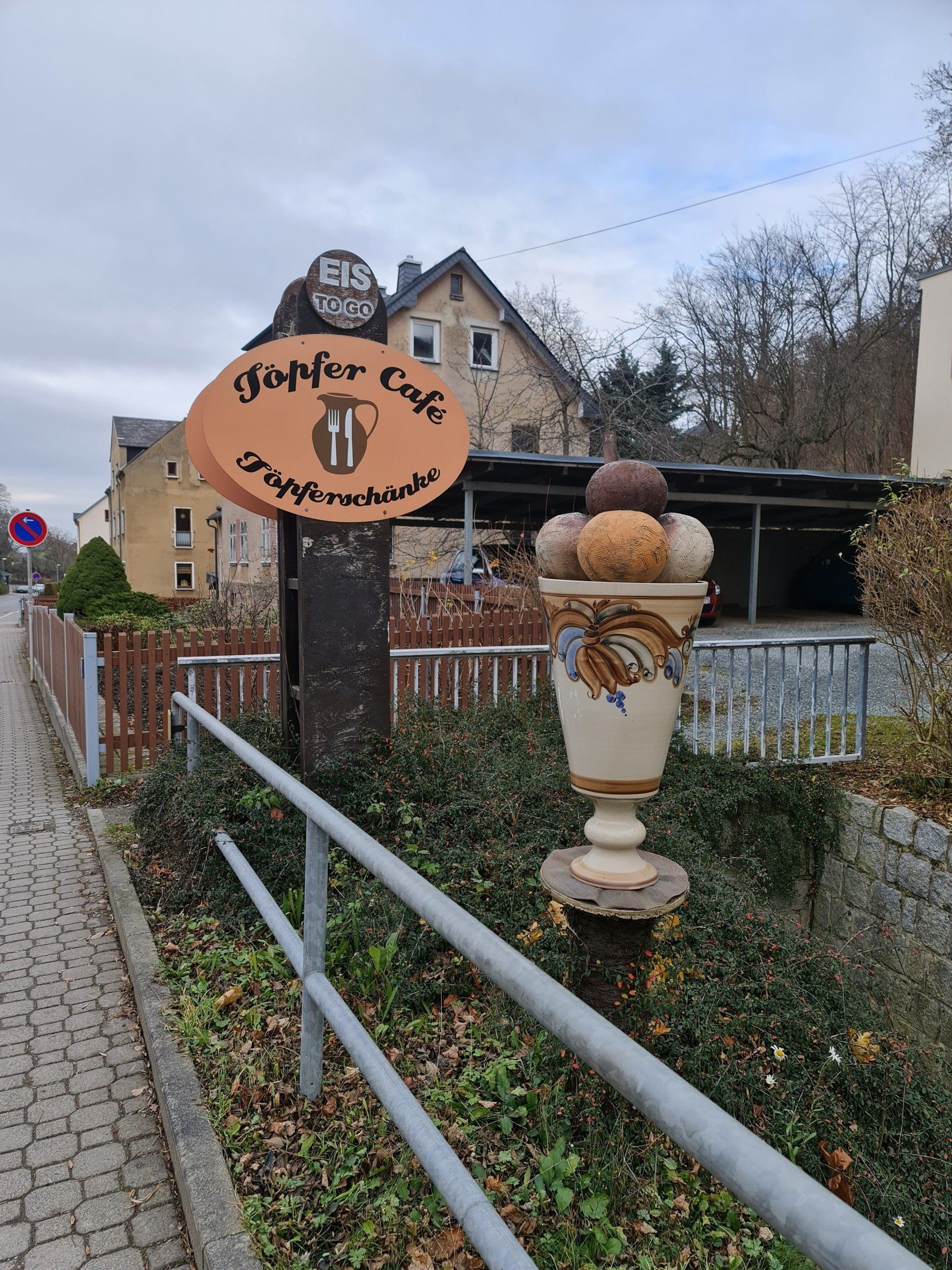 Hinter dem grauen Geländer ein großer Eisbecher aus Keramik und ein Schild, dass auf die Töpferei und das Café hinweist