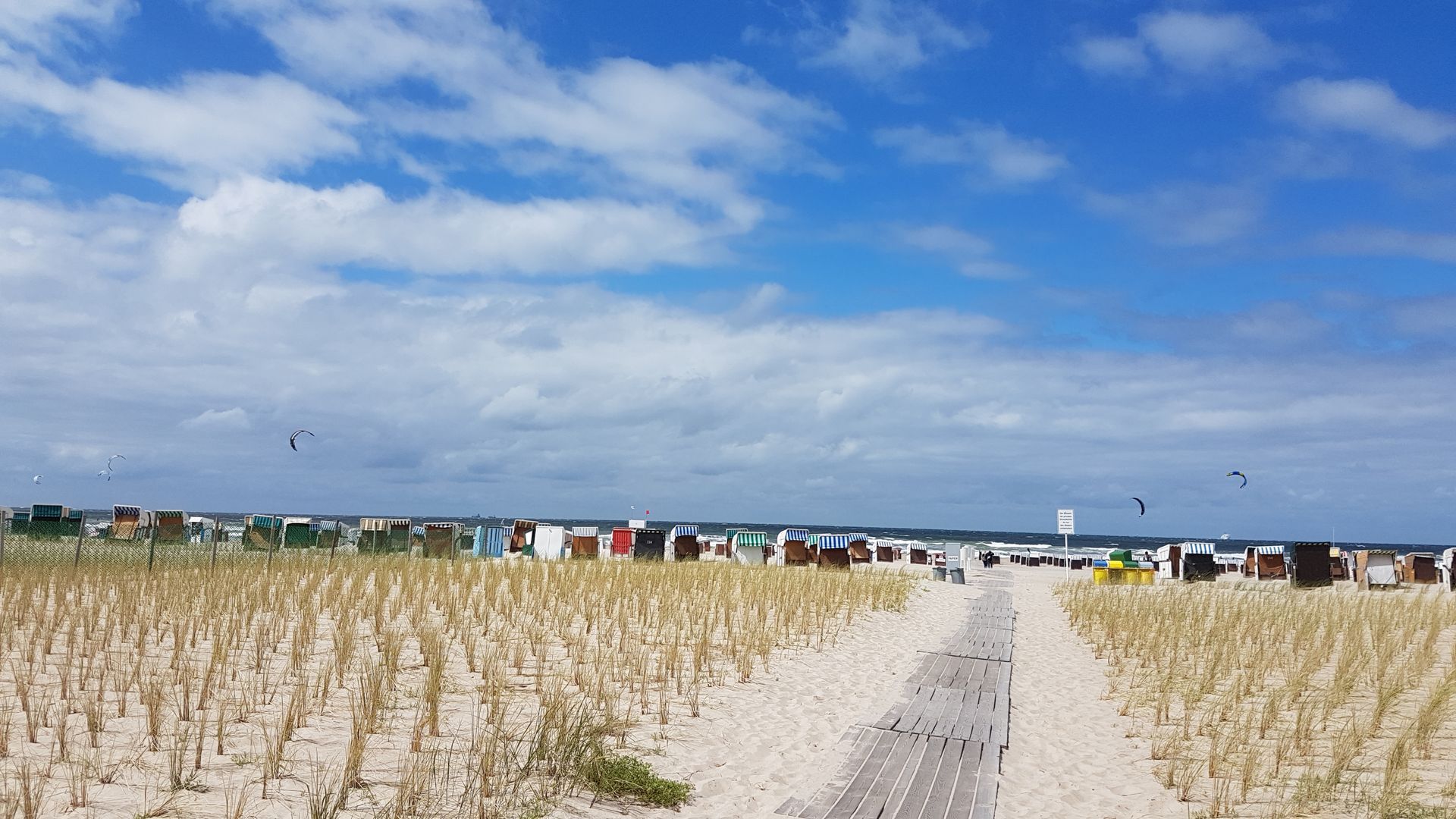 blauer Himmel mit kleinen Wölkchen ein Pfad aus Brettern verläuft über einen breiten Sandstrand. Der Strand ist mit trockenen Gräsern bepflanzt.