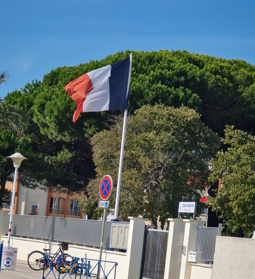 große Pinien im Hintergrund, davor Zaun und die Flagge von Frankreich weht
