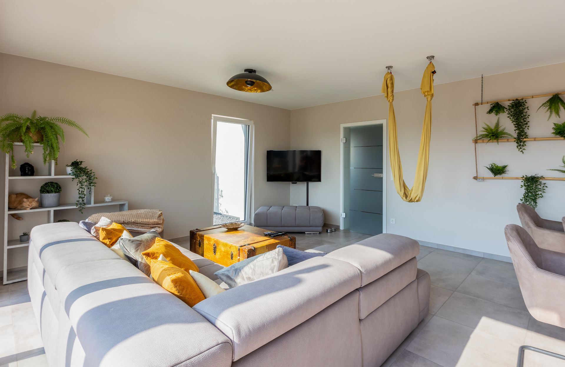 Eine schwarz goldene Deckenlampe hängt über einem Truhentisch. Der Tisch steht vor einem gräumigem grauem Ecksofa, welches mit reichlich Kissen in den Farben sonnengelb, grau und weiß belegt ist. Rechts im Bild hängt ein gelbgoldenes Yogatuch.