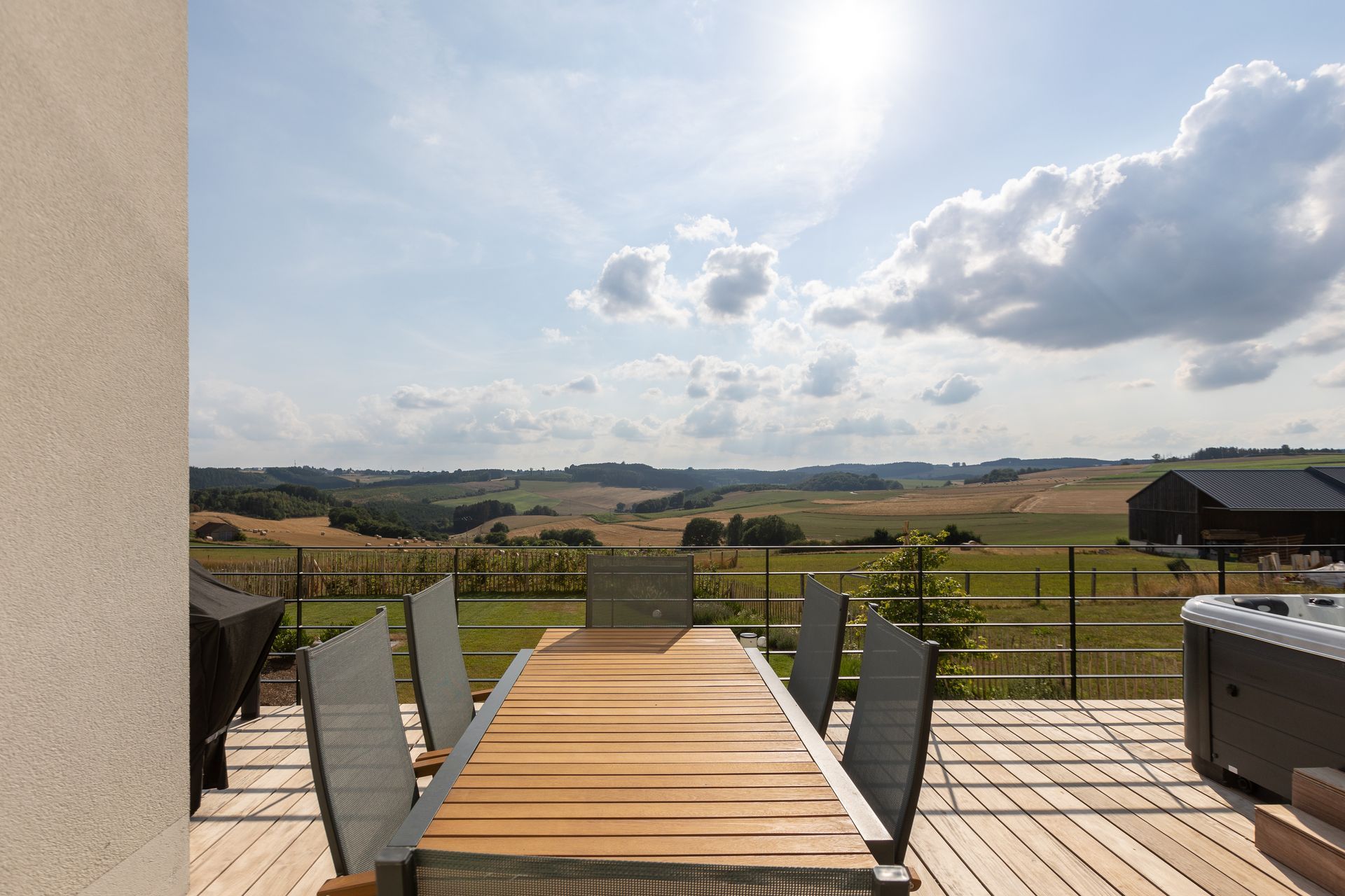 Hellblauer Himmel mit ein paar hellen Wolken. Die Sonne scheint über das grüne Tal. Im Vordergrund sieht man den Blick aus der Terrassentür. Dort steht ein brauner Holztisch mit 5 grauen Stühlen.