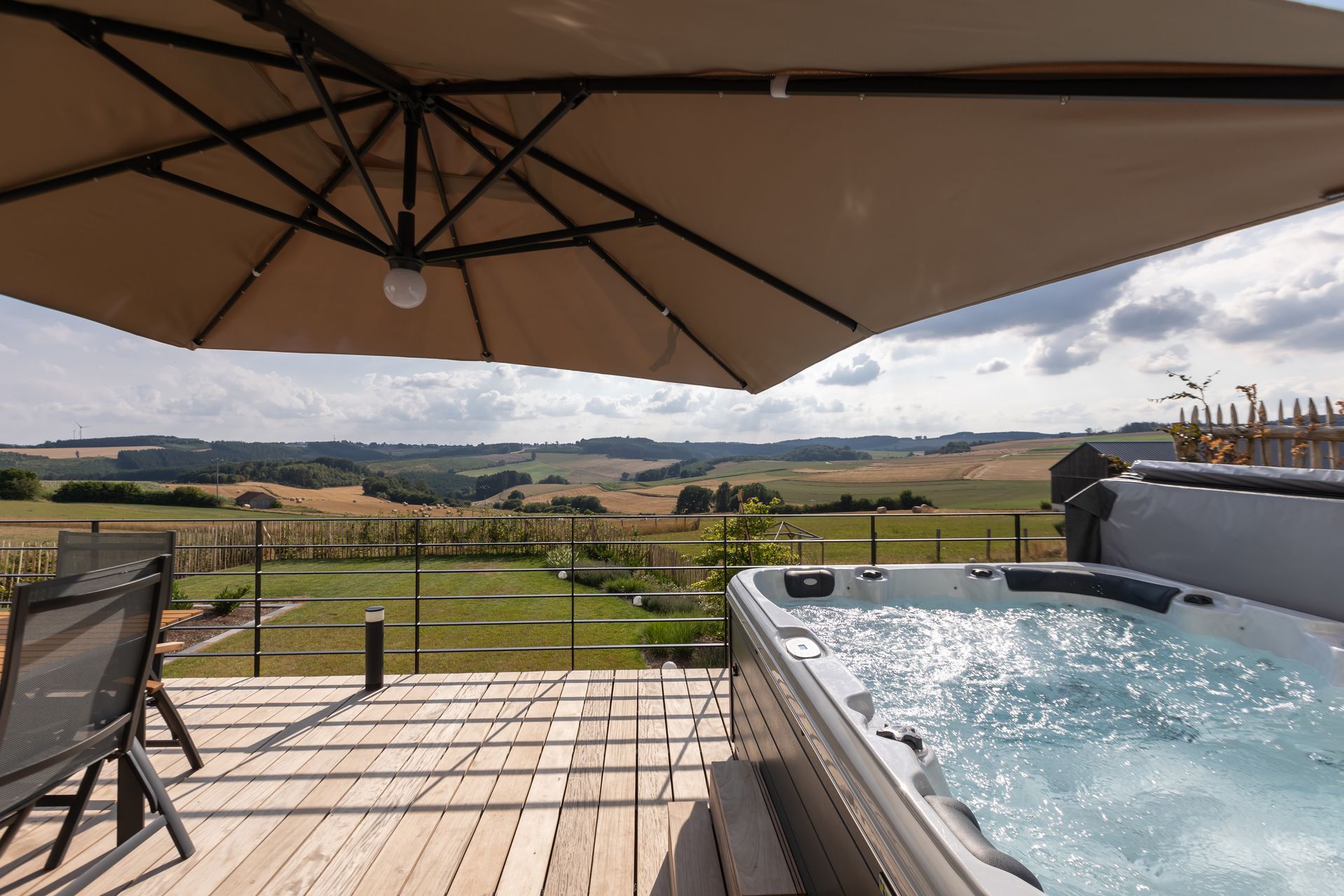 Hellblauer Himmel mit ein paar hellen Wolken. Die Sonne scheint über das grüne Tal. Im Vordergrund sieht man den Jacuzzi. Der beige Sonnenschrim ist aufgespannt. Die Terrasse ist eingezäunt mit einem anthrazitfarbenem Geländer.