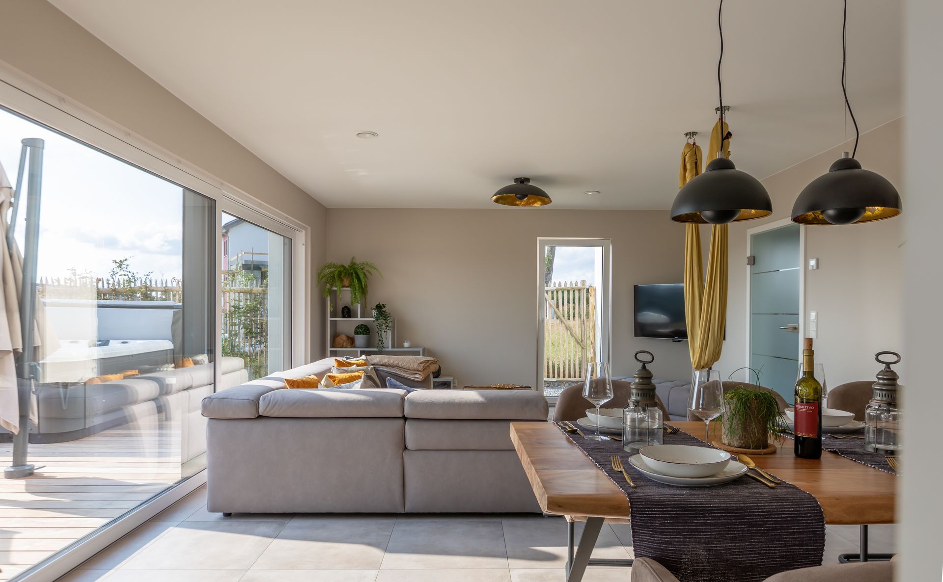 Das graue Ecksofa ist seitlich zu sehen. Im vorderen rechten Bereich des Bildes ist ein Stück des Tisches sowie ein Sthul zu sehen, um einen Eindruck des Wohnbereiches gewinnen zu können.