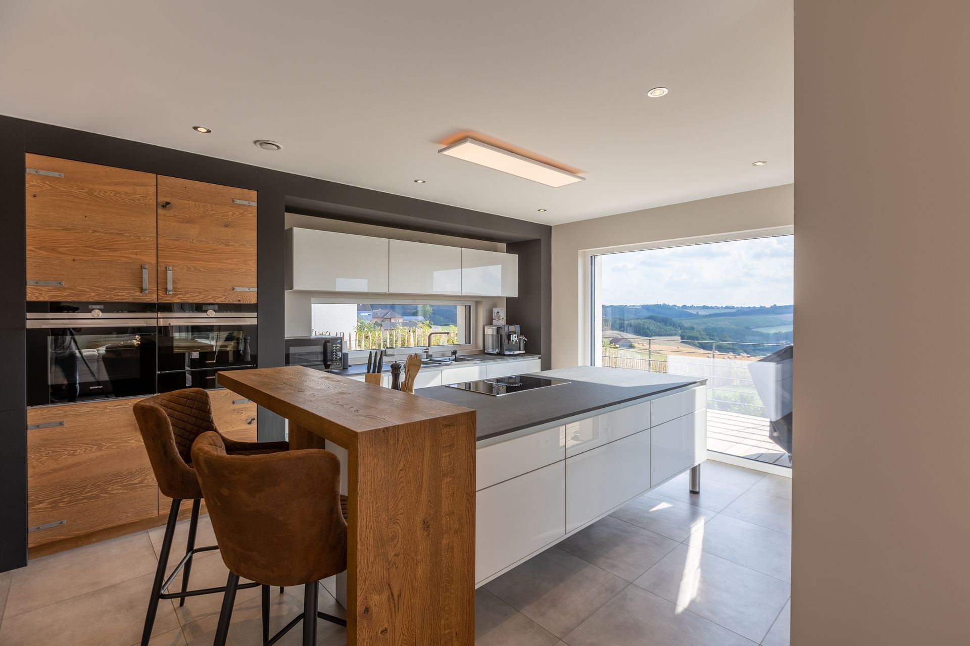 Die große weiße Kochinsel ist in der Mitte des Bildes zu sehen. Links der Insel stehen zwei braune Barhocker, welche weitere Sitzmöglichkeiten bieten. An der schwarz gestrichene Wand befinden sich weiße Hängeschränke.