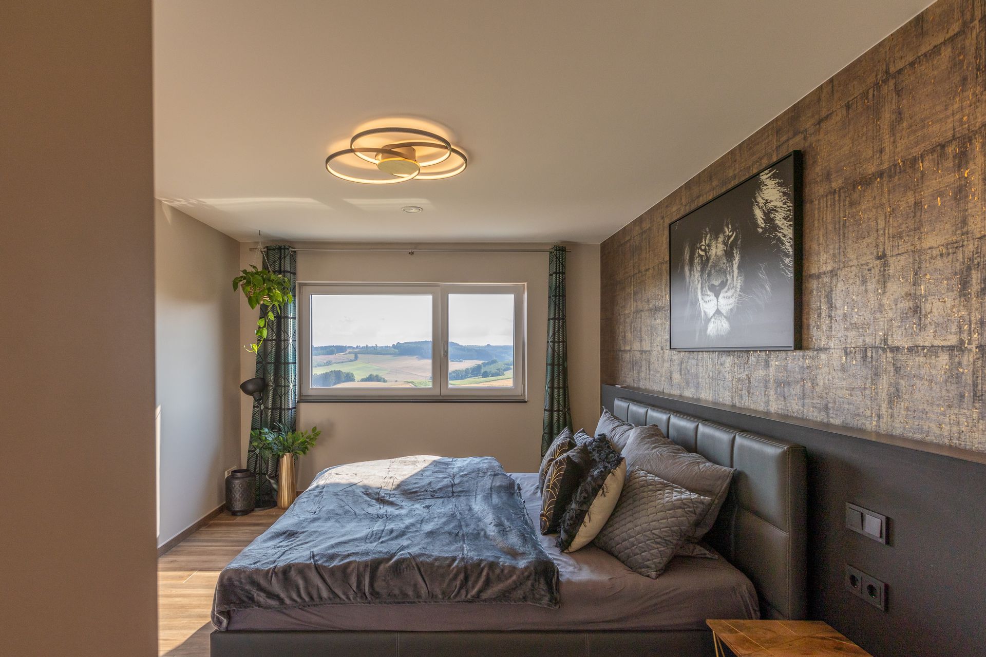 Das graue Tempur Bett ist mit grauer Bettwäsche und einer grauen Tagesdecke bezogen. Der Blick führt in den Hauseigenen Garten. Eine Hängepflanze ragt aus einem goldenem Blumentopf von der Decke, welcher sich in einer Blumenampel befindet. Das gäserne Löwenbild hängt an einer goldenen Wand über dem Bett.