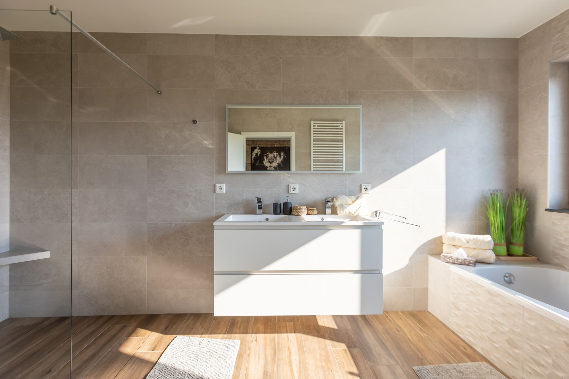 Hinter einem Spiegel, welcher beleuchtet werden kann, sind große hellgraue Fliesen. Unter dem Spiegel hängt ein Doppelwaschbecken, welches in einem weißen Unterschrank eingefasst ist. Der Boden ist in Holzoptik gefliest.