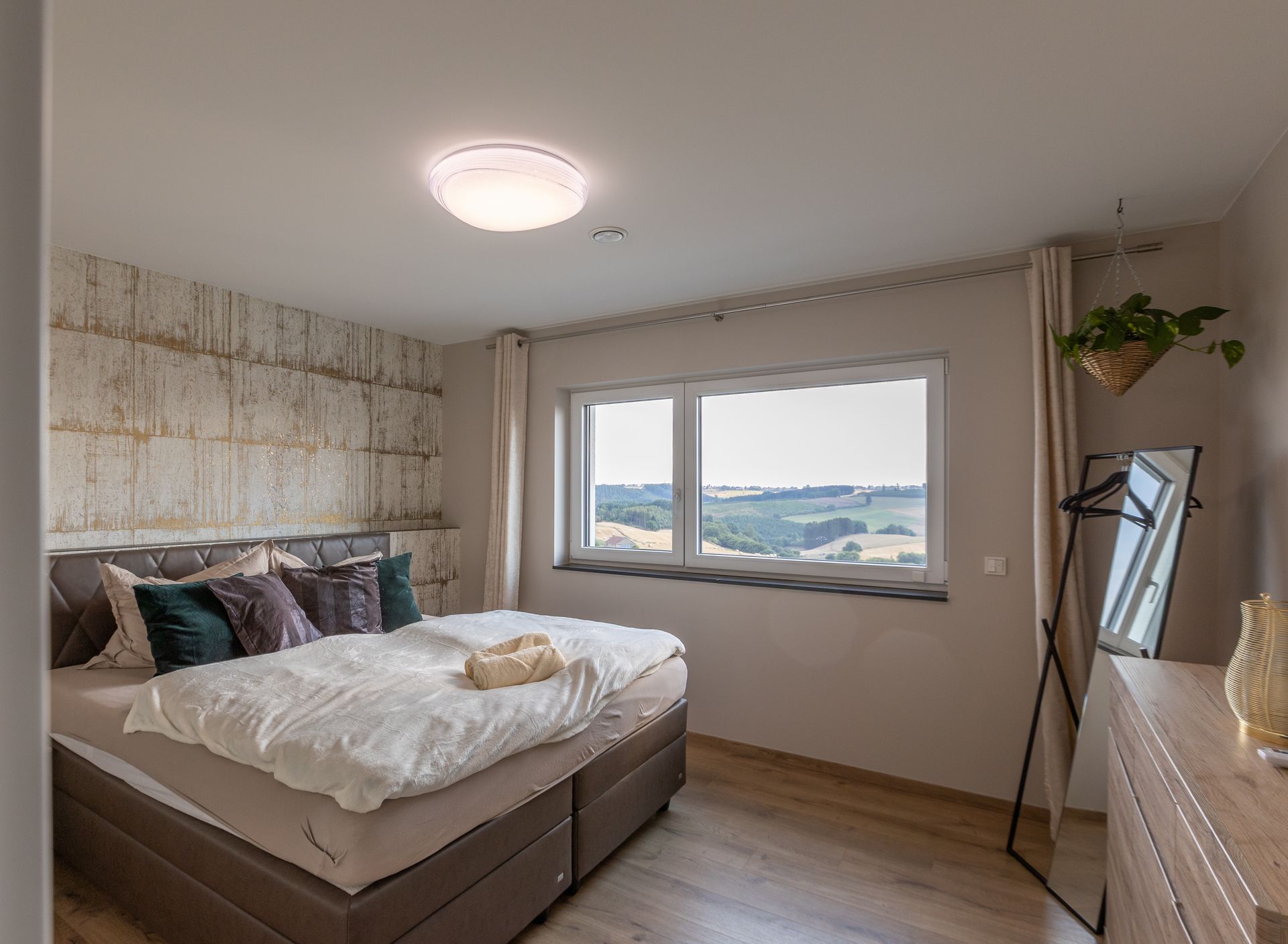 Ein braunes Boxspringbett steht vor einer weiß goldenen Wand. DIe beige Tagesdecke liegt vor den schwarz goldenen Kissen. Auf der Decke liegen zwei beige Handtücher, welche gerollt positioniert wurden.
