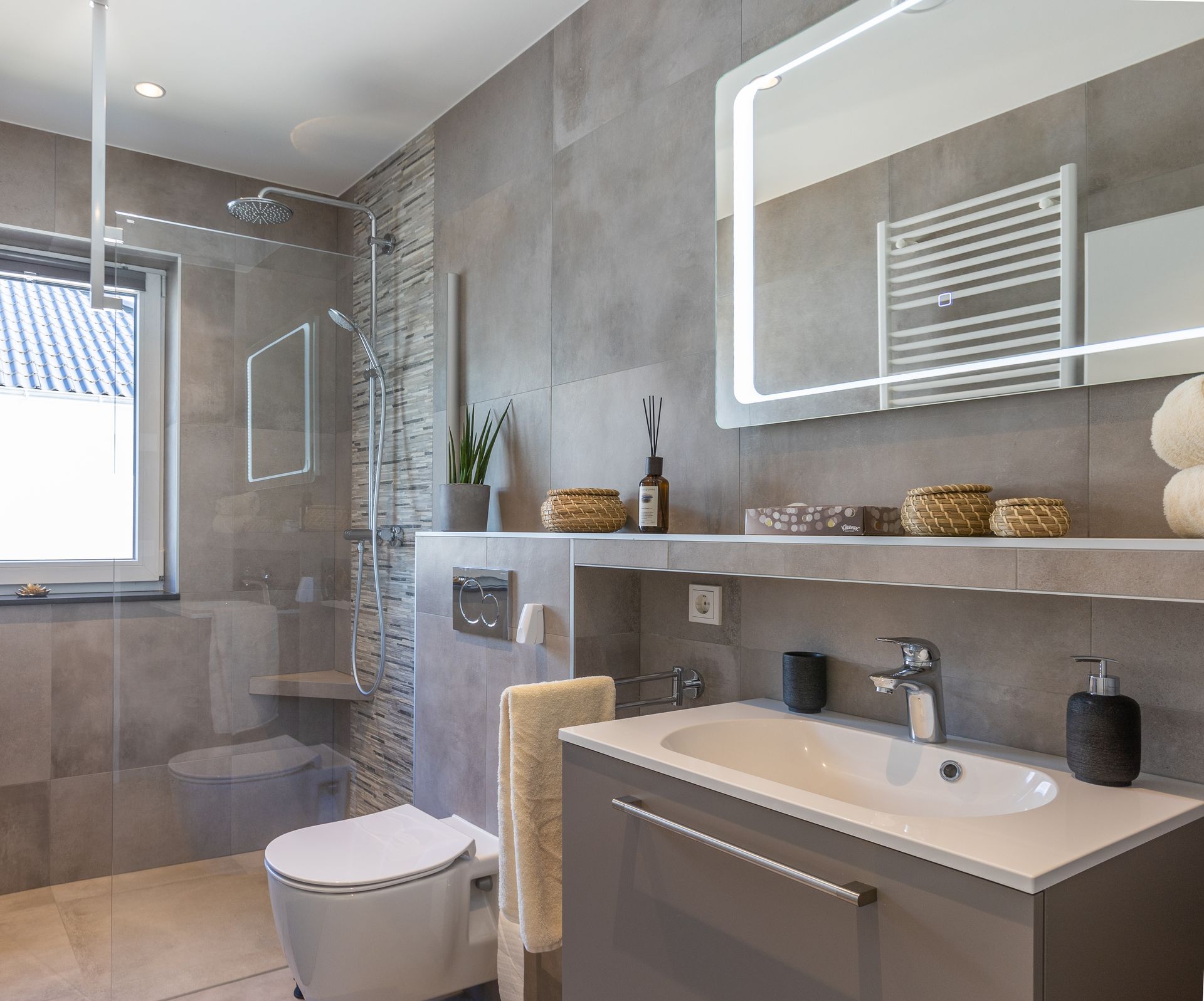 Ein graues Badezimmer mit einer Dusche (welche mit einer Italienischen Wand begrenzt ist, befindet sich hinter dem Fenster. Vor dieser Wand ängt eine weiße Toilette. Der Waschbeckenunterchrank rechts im Bild ist grau mit einem silbernem Griff. Kleine Bastkörbchen stehen auf der Ablage über dem Waschbecken. Ein beleuchteter Spiegel hängt über dem Waschbecken.