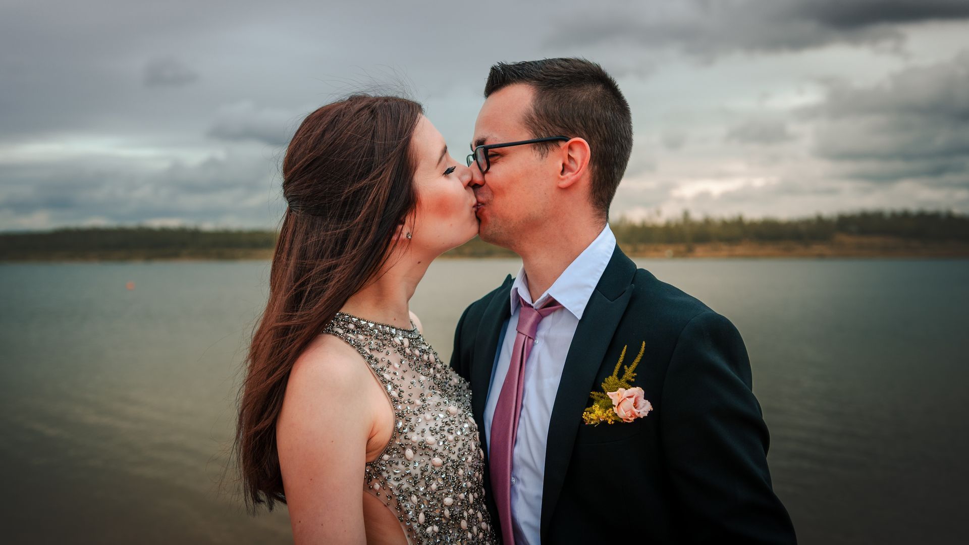 Romantisches Hochzeitsfoto am See – Brautpaar küsst sich bei windigem Wetter, Braut im glitzernden Kleid und Bräutigam im schwarzen Anzug mit rosa Krawatte