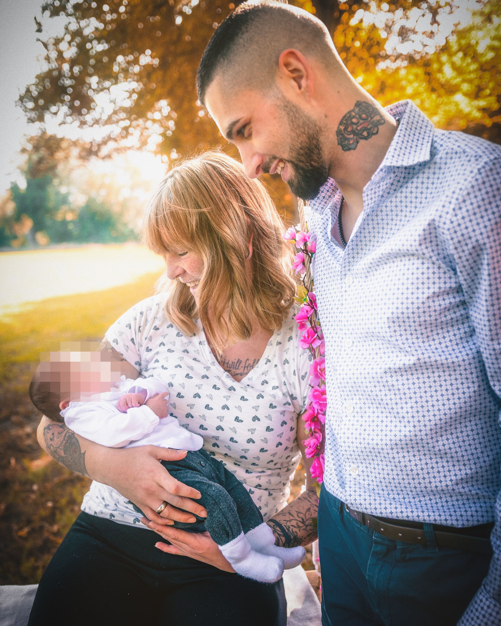 Mutter hält ihr Neugeborenes liebevoll im Arm, während der Vater sich von der Seite lächelnd dazuneigt. Die Familie steht in warmem Herbstlicht unter einem blühenden Ast mit rosa Blumen – eine ruhige, innige Szene voller Nähe.
