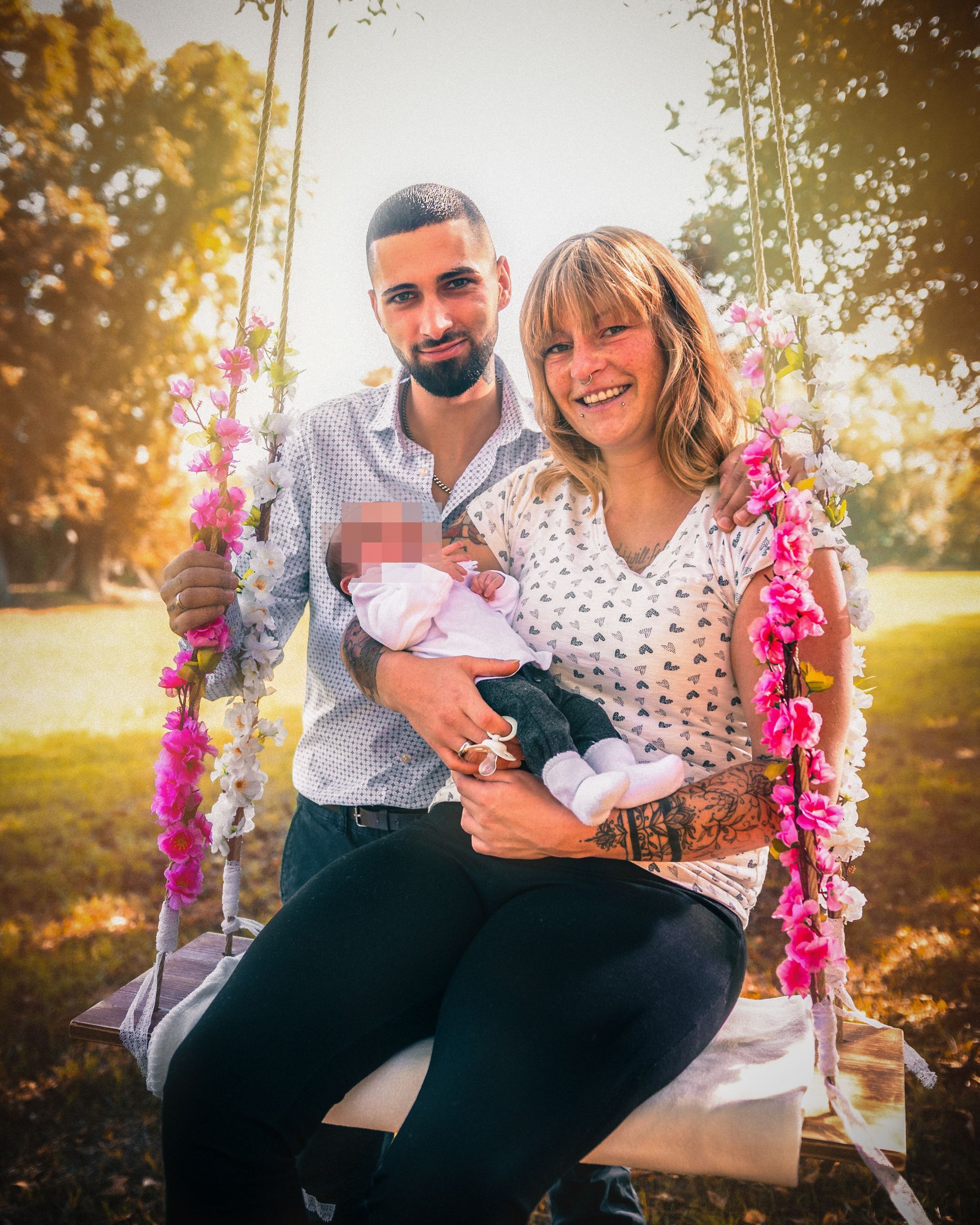 Junge Familie mit Neugeborenem sitzt auf einer mit Blumen geschmückten Holzschaukel im Park. Die Eltern lächeln in die Kamera, das Baby liegt schlafend auf dem Arm der Mutter. Natürliches Licht, herbstliche Stimmung.
