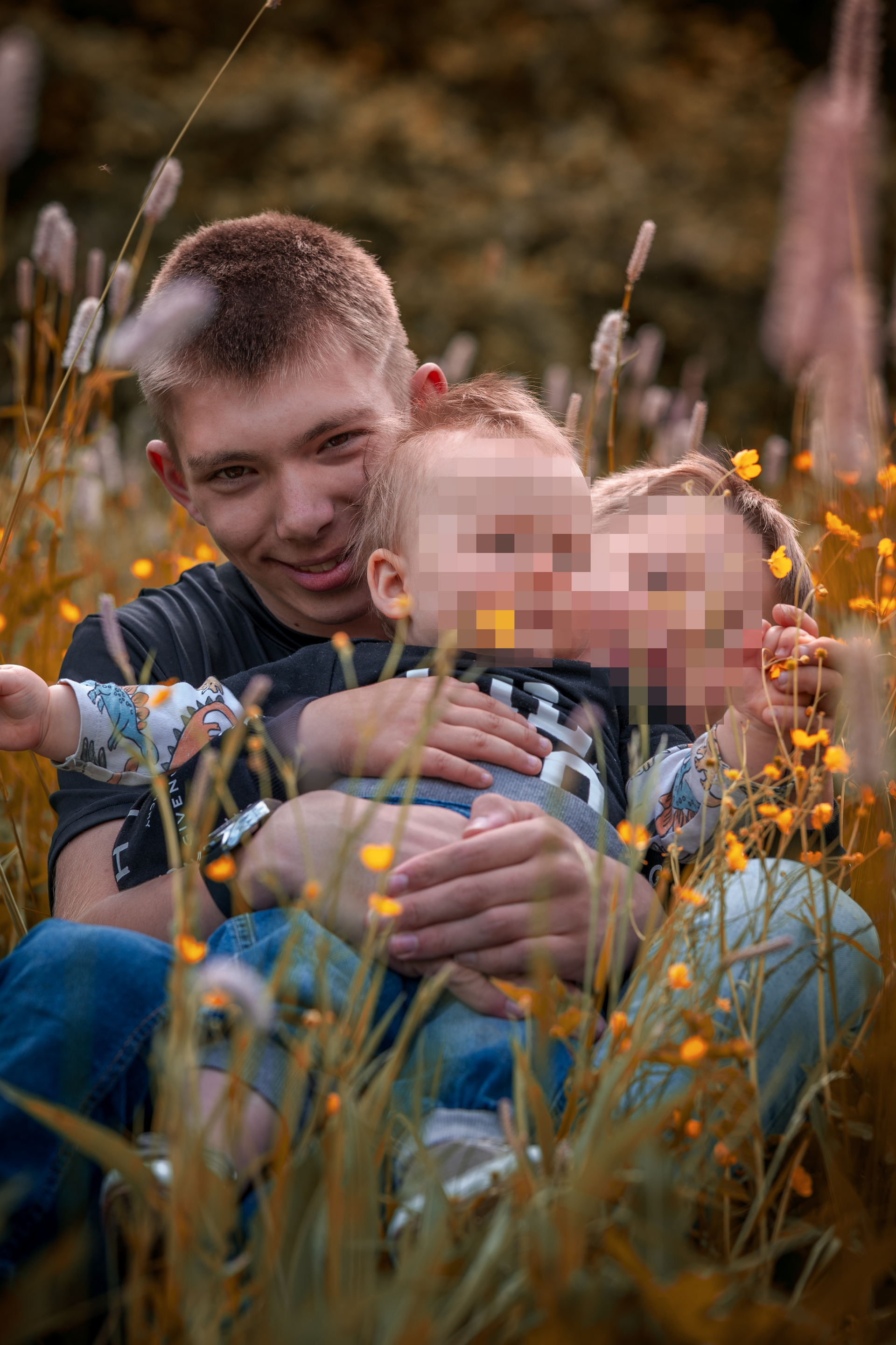 Großer Bruder umarmt zwei kleine Geschwister im hohen Gras mit gelben Wildblumen – authentisches Familienshooting auf einer Sommerwiese