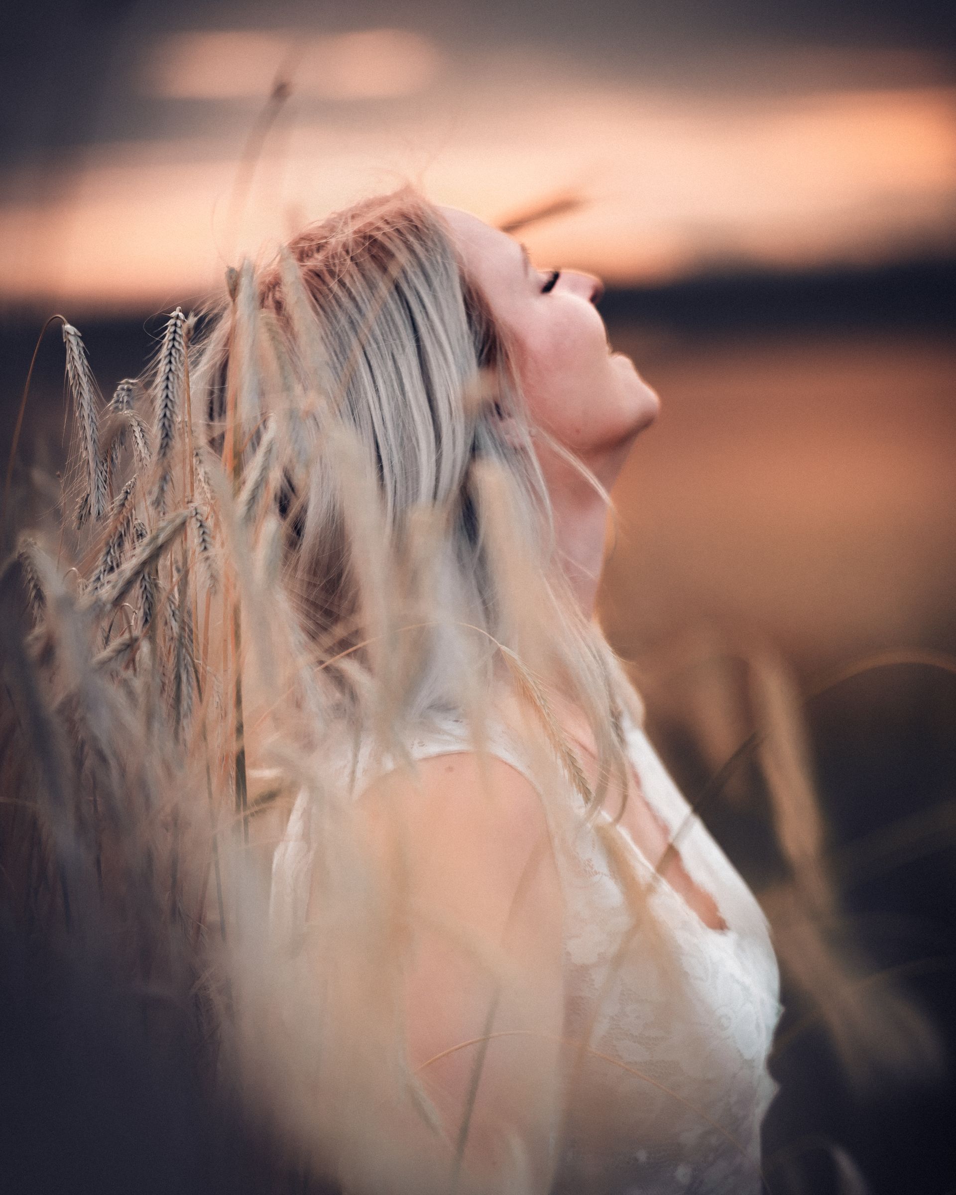 Frau mit blondem Haar steht mit geschlossenen Augen in einem Kornfeld bei Sonnenuntergang. Ihr Gesicht ist dem Licht zugewandt, sie wirkt ruhig, gelöst und eins mit der Natur. Die goldenen Halme umrahmen ihr Profil sanft.