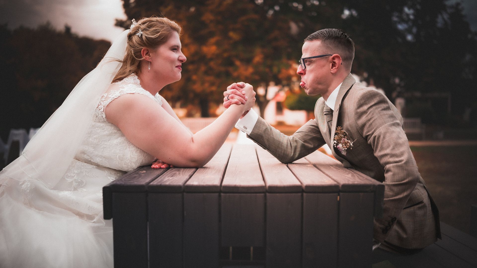 Braut und Bräutigam liefern sich beim Armdrücken ein humorvolles Duell an einem Holztisch im Freien während ihrer Hochzeit auf dem Gelände des Kloster Nimbschen bei Grimma.
