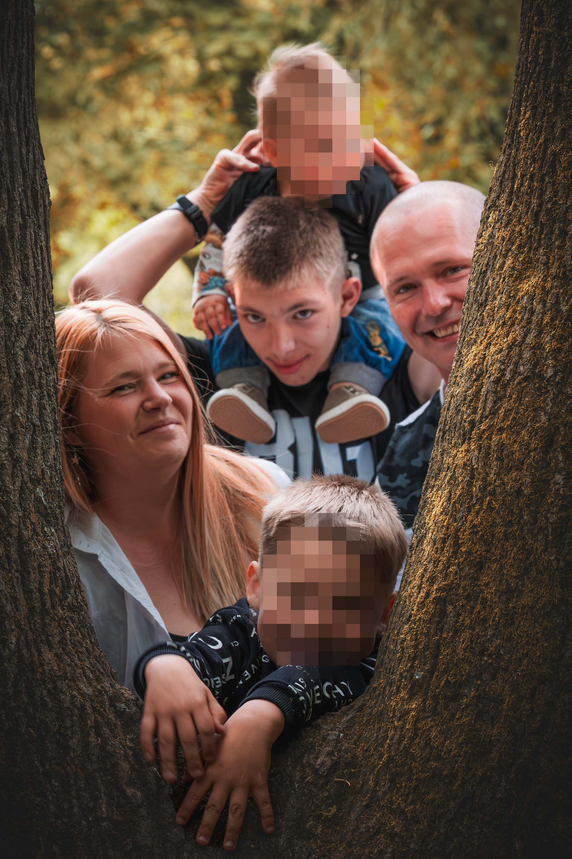 Fröhliche Familie mit drei Kindern posiert lachend zwischen zwei Baumstämmen in einem herbstlichen Park – natürliches Outdoor-Familienshooting