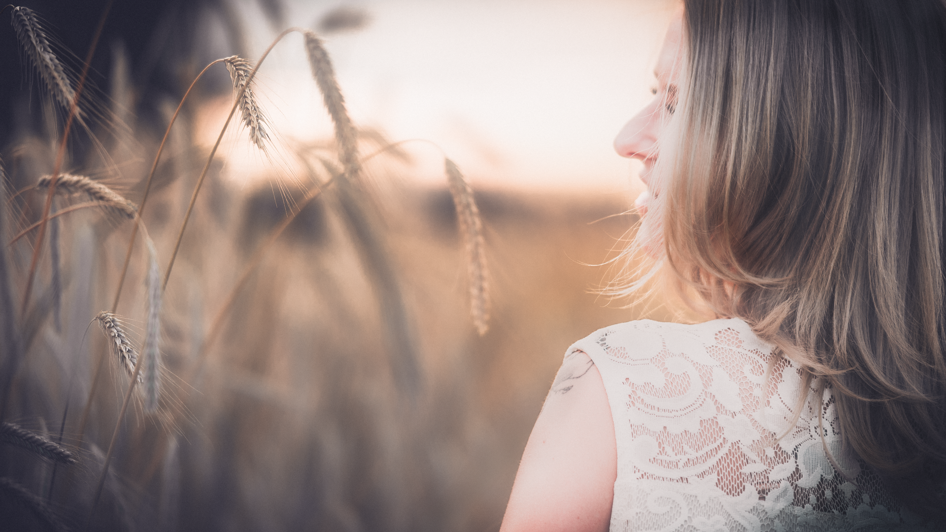 Frau mit blondem Haar steht im Kornfeld und blickt bei Sonnenuntergang nach rechts. Der Fokus liegt
