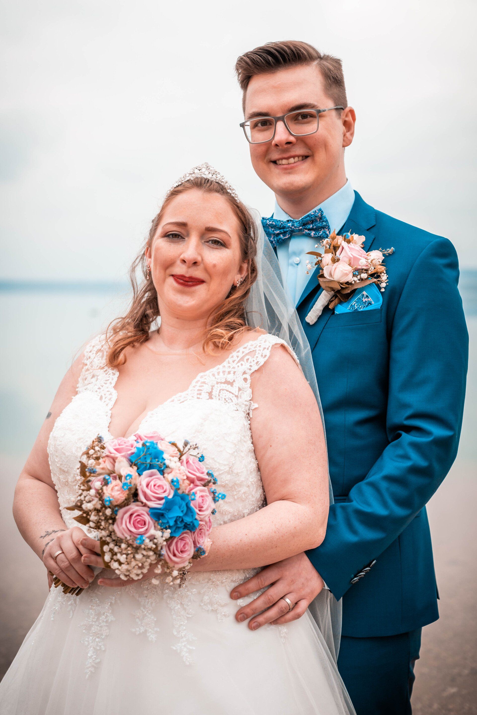 Klassisches Hochzeitsfoto am Wasser – Braut mit Spitzenkleid und bunter Rosen-Brautstrauß, Bräutigam im blauen Anzug umarmt sie liebevoll von hinten