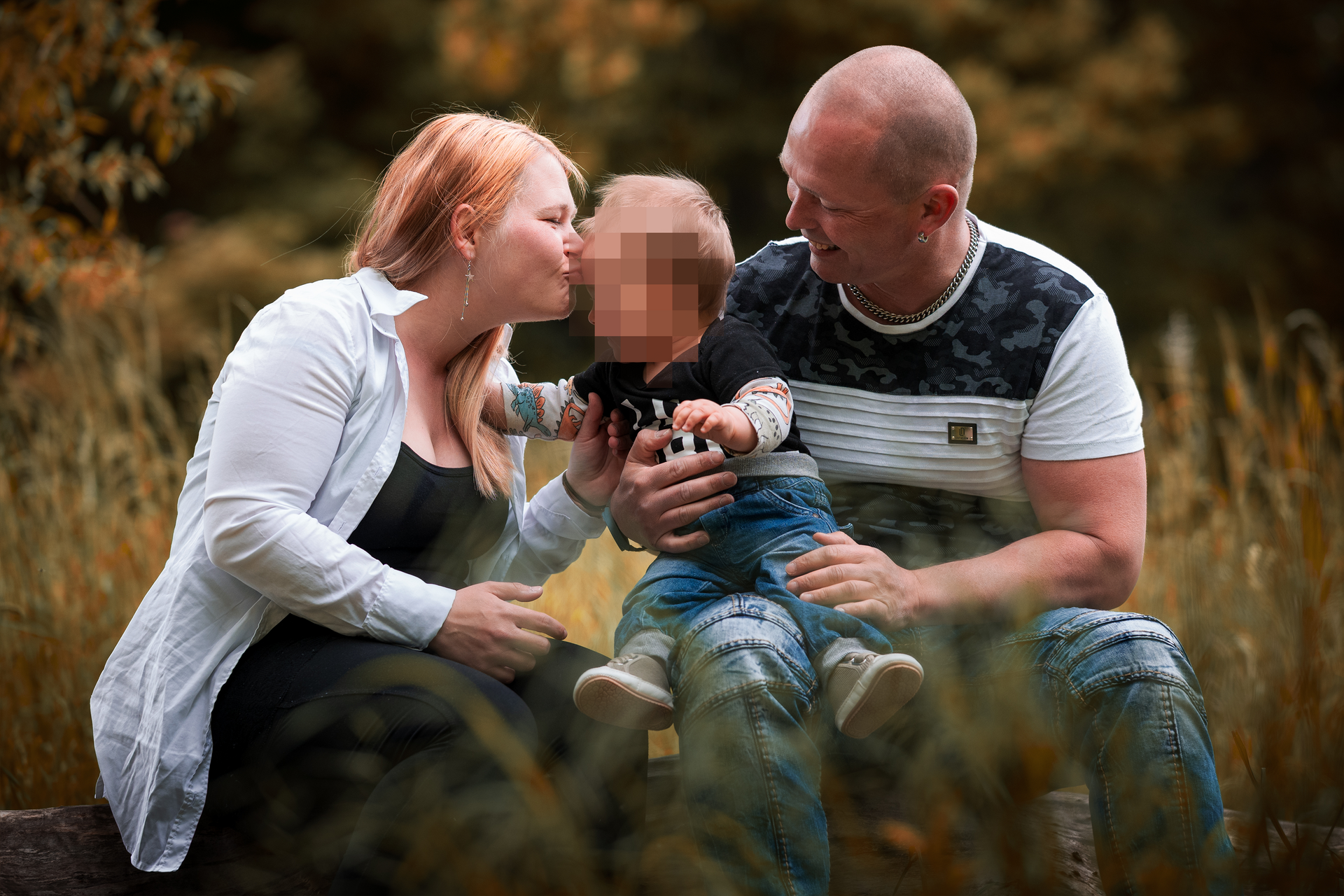 Lächelnde Eltern sitzen mit ihrem Kleinkind auf einem Baumstamm inmitten herbstlicher Natur