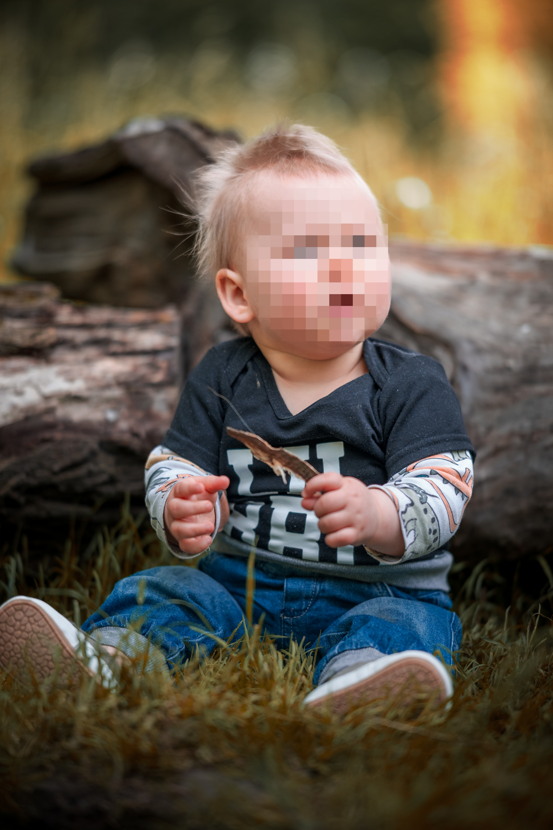 Kleiner Junge im T-Shirt mit der Aufschrift „LIL BRO“ sitzt auf einer Wiese vor Baumstämmen und hält einen Ast – natürliches Babyshooting im Freien