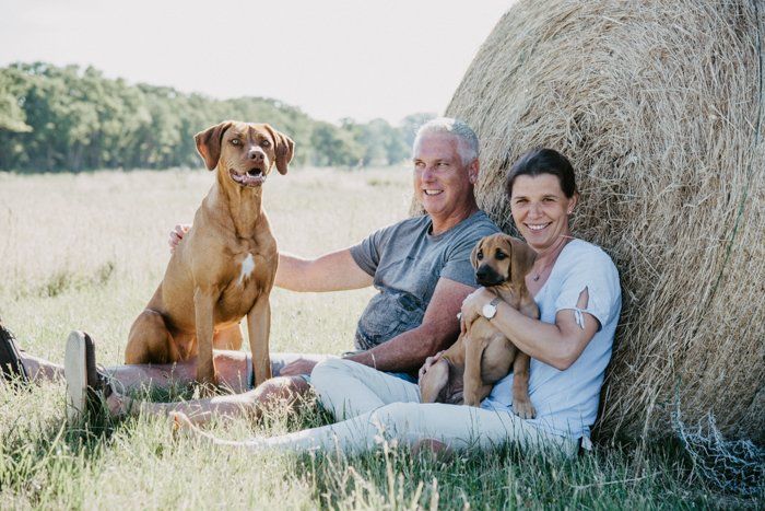 <img src=”Paar-mit-Hunden-auf-Wiese.jpg” alt=”Ein Paar sitzt mit ihren zwei Hunden auf einer Wiese vor einem Rundballen”>