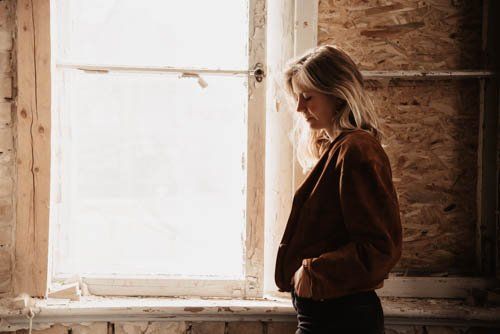 <img src=”junge-Frau-steht-an-altem-Fenster.jpg” alt=”Eine junge Frau steht an einem alten Fenster und schaut zu Boden”>