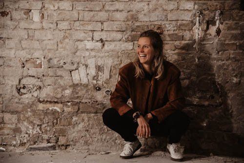 <img src=”junge-Frau-hockt-vor-alter-Mauer-lacht.jpg” alt=”Eine junge Frau hockt lachend vor einer alten Mauer”>