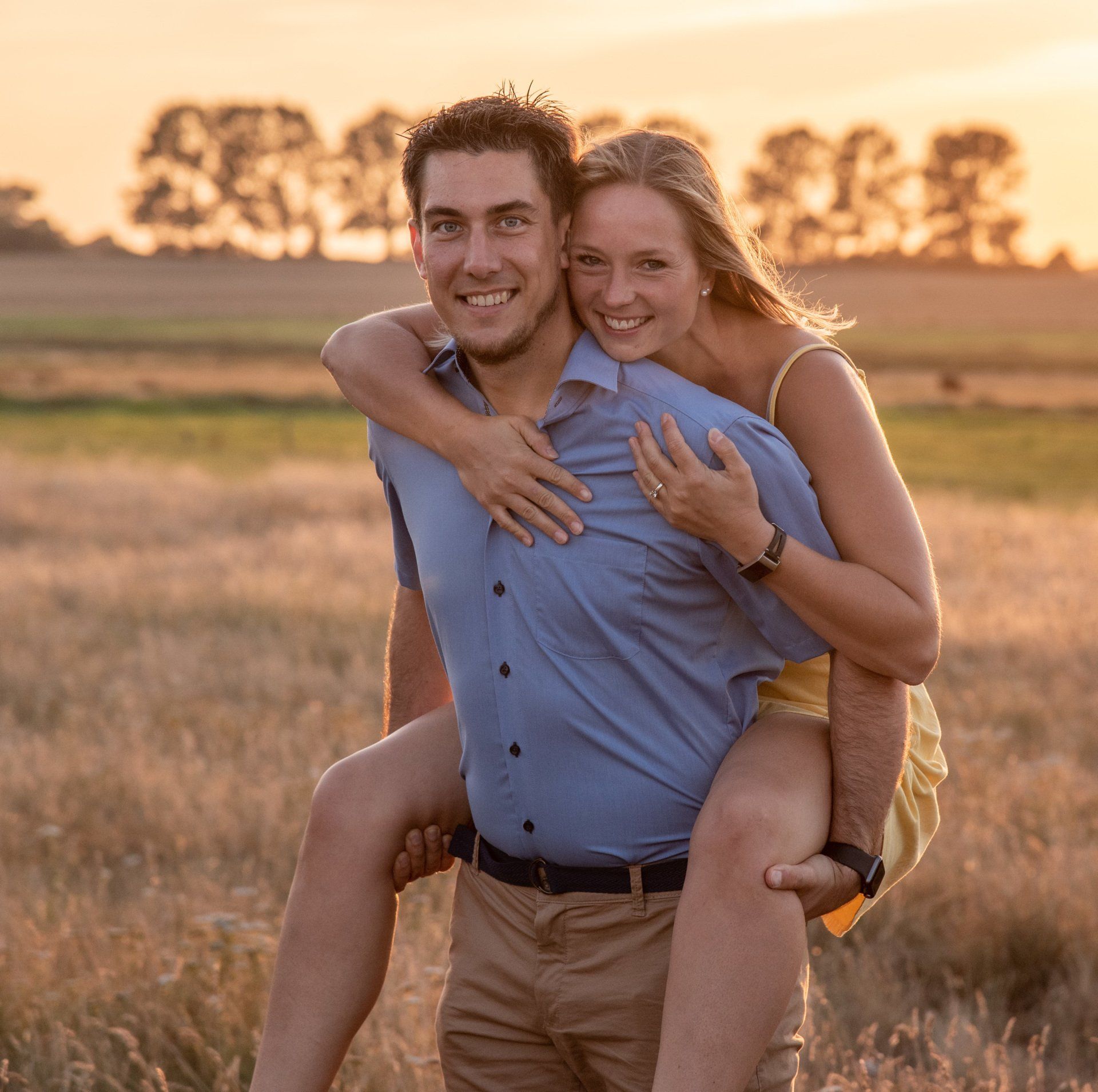 <img src=”junge-Frau-auf-huckepack-im-Sonnenuntergang-lachend.jpg” alt=”er trägt sie huckepack auf einer Wiese im Sonnenuntergang”>