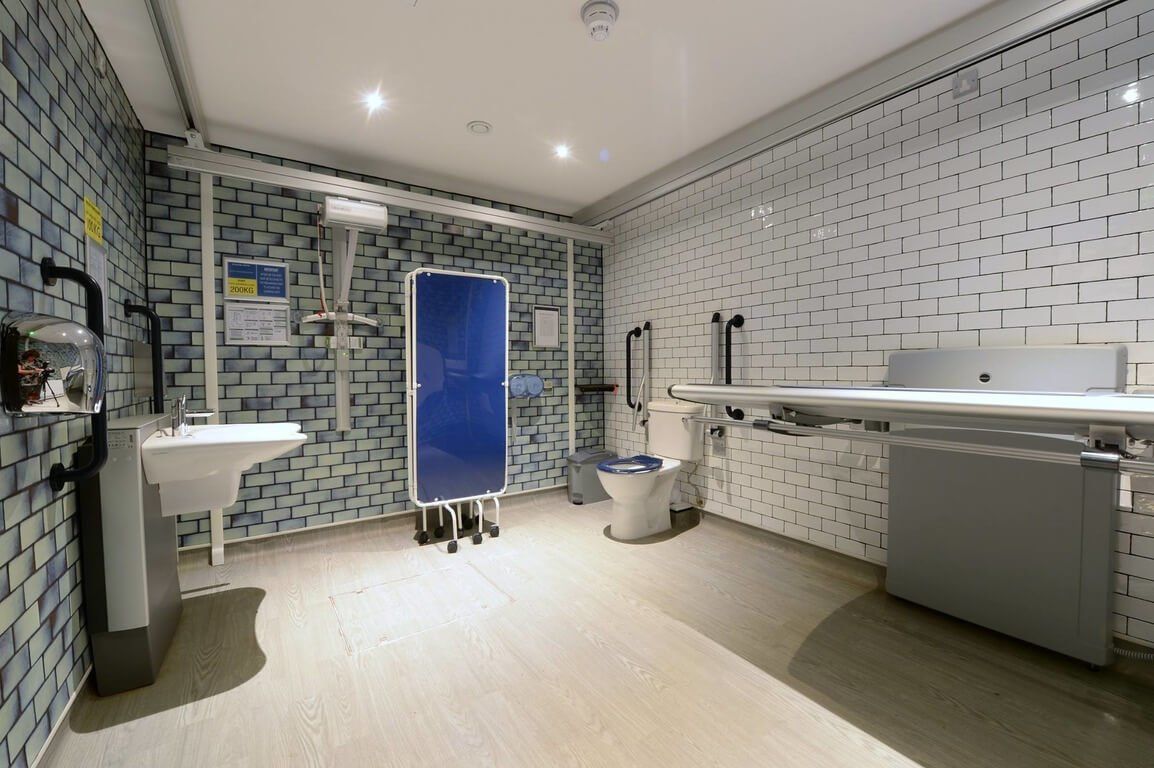 a changing places toilet showing a old fashioned tiled wall wall changing bed on the right hand side with a changing bed sink on the left hand side and a toilet in the middle of the room at the back