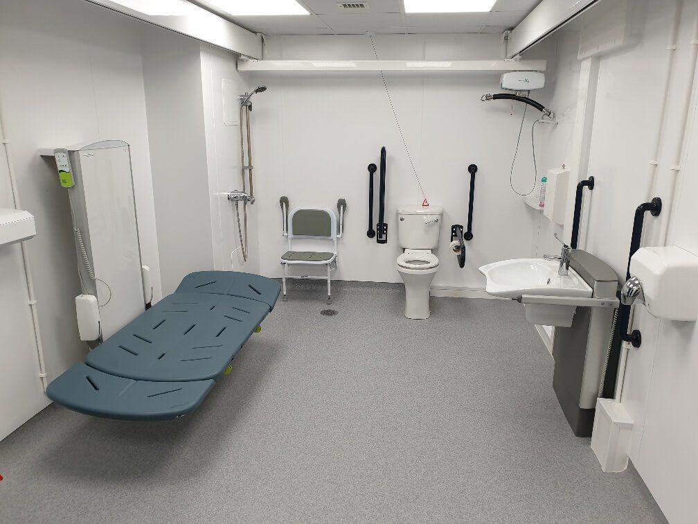 a clinical looking changing places toilet with a changing bed  on the left hand side, toilet against the back wall and a sink on the right hand side