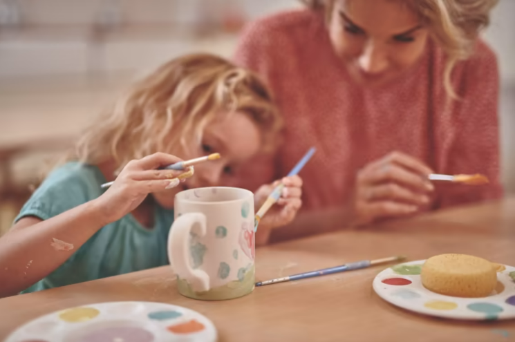 an image of a young girl painting a mug at the table with her mother