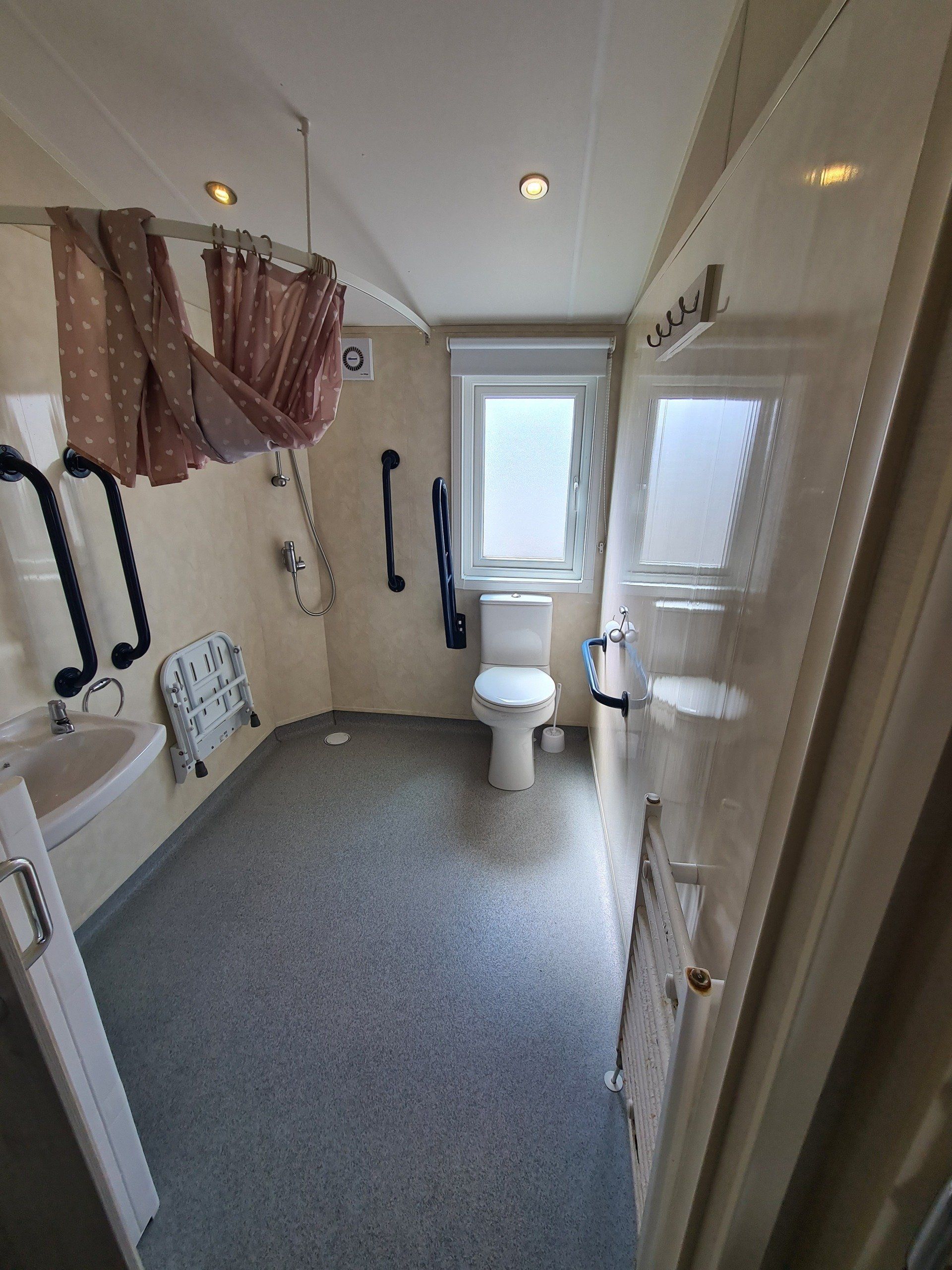 a picture of the wetroom from the hallway showing the toilet with grab handles, shower in the corner and a foldable seat under the shower
