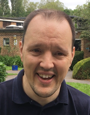 A smiling man with buildings and bushes in the background