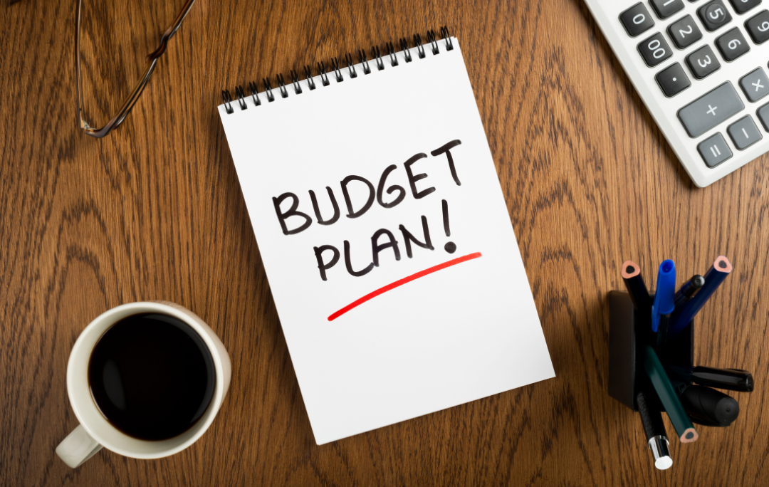 Wooden desk with coffee, glasses, pencils and calculator, and a notepad saying Budget Plan!