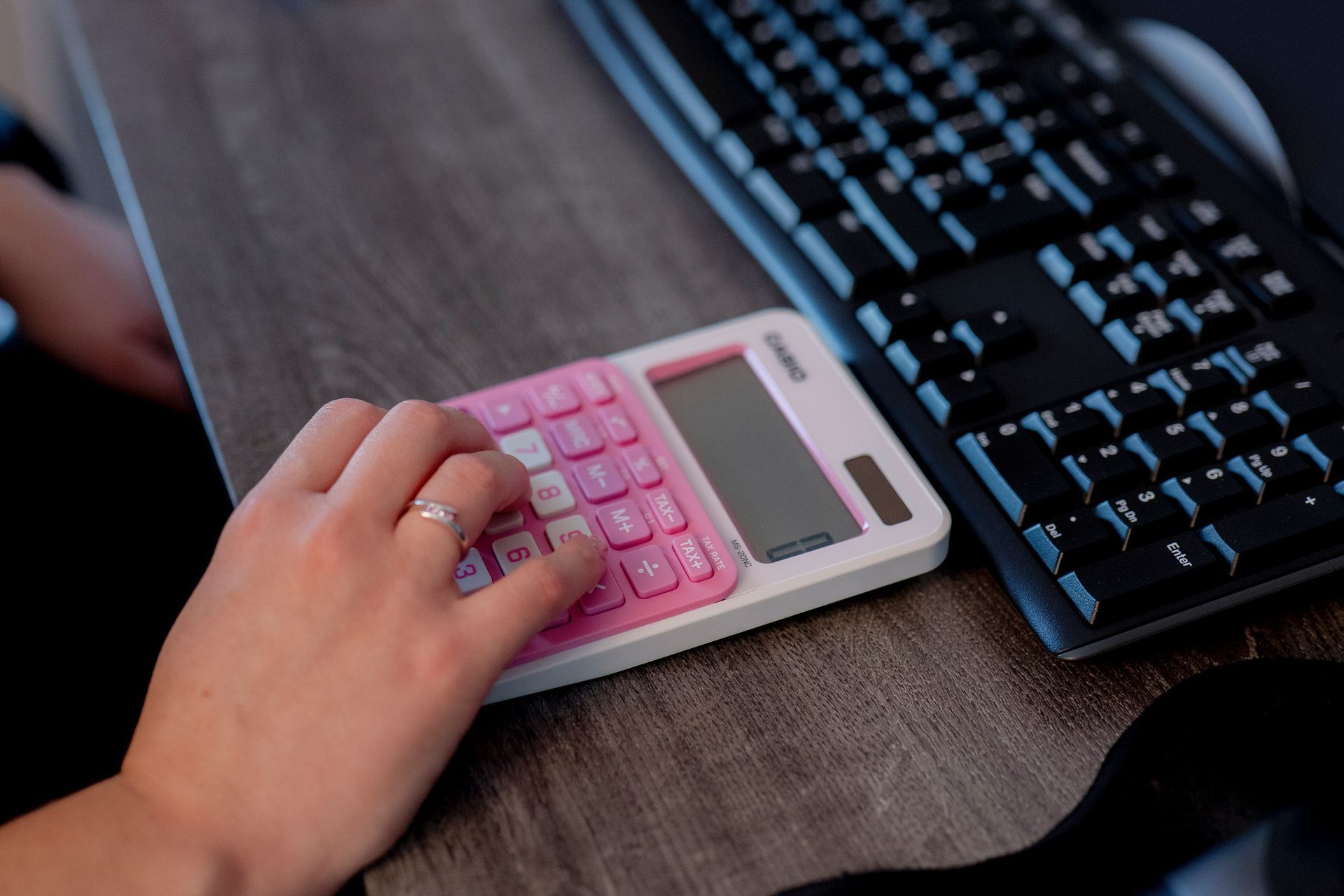 A hand using a pink calculator on a desk with a keyboard
