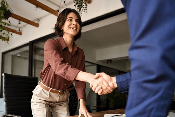 Two people in casual business attire smiling and shaking hands in a relaxed, friendly setting.