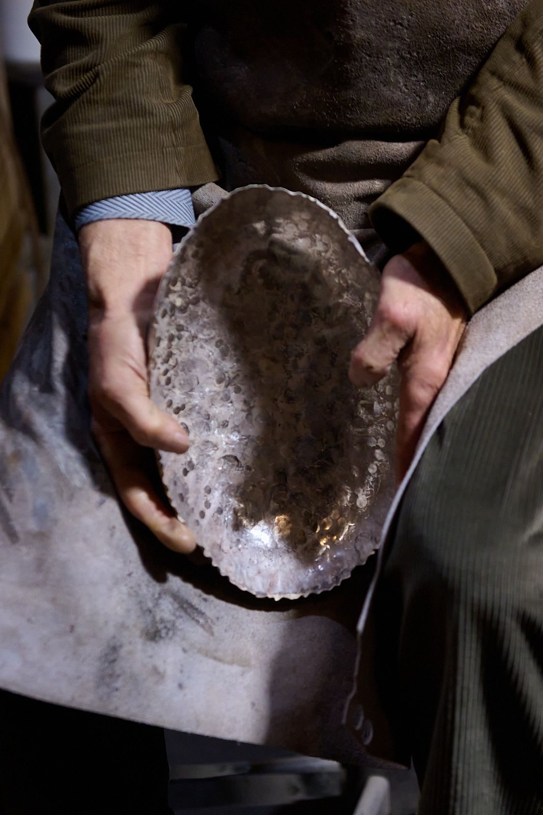 main d'artisan agé tenant un bol en argent entre ses genoux devant un tablier en cuir élimé