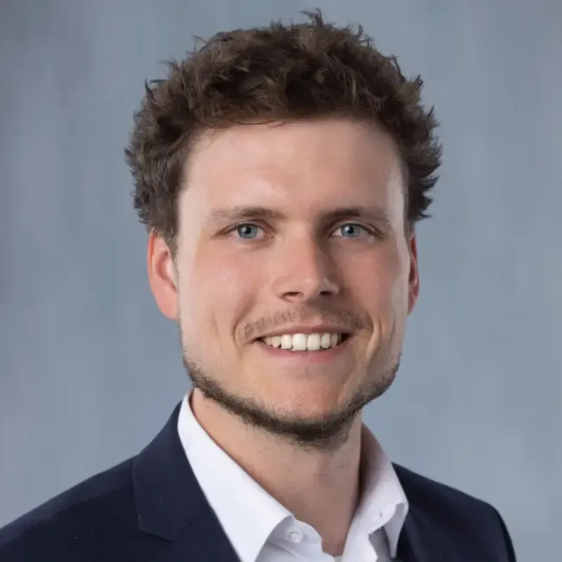 Porträt von Dr.-Ing. Sebastian-Paul Kopp, Geschäftsführer des Bayerischen Laserzentrums, lächelnd vor grauem Hintergrund.
