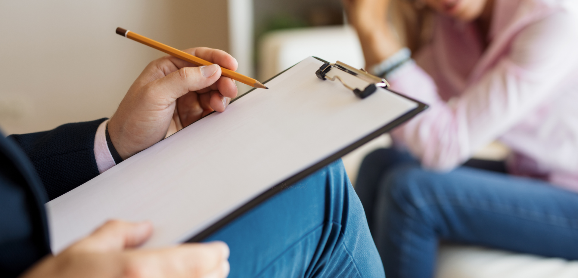 zu sehen ist ein Bein eines Mannes, der Mann scheint zu sitzen und hat auf dem Schoß eine Kladde liegen, darauf ein leeres weißes Blatt Papier. Er hat einen Stift in der Hand und ist bereit etwas zu schreiben. Im HIntergund sitzt auf einer Couch eine Frau mit Jeanshose und Rosa Pullover. Man sieht von Ihr nur den Rumpf und die Oberschenkel.