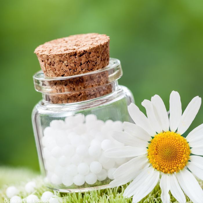 vor einem Grasgrünen Hintergrund ist ein kleines Gefäß mit weißen kleinen Kügelchen zu sehen, das Gefäß ist mit einem Korkdeckel verschlossen. Recht im Bild etwas vor der Flasche ist die Blüte eines Gänseblümchens zu sehen