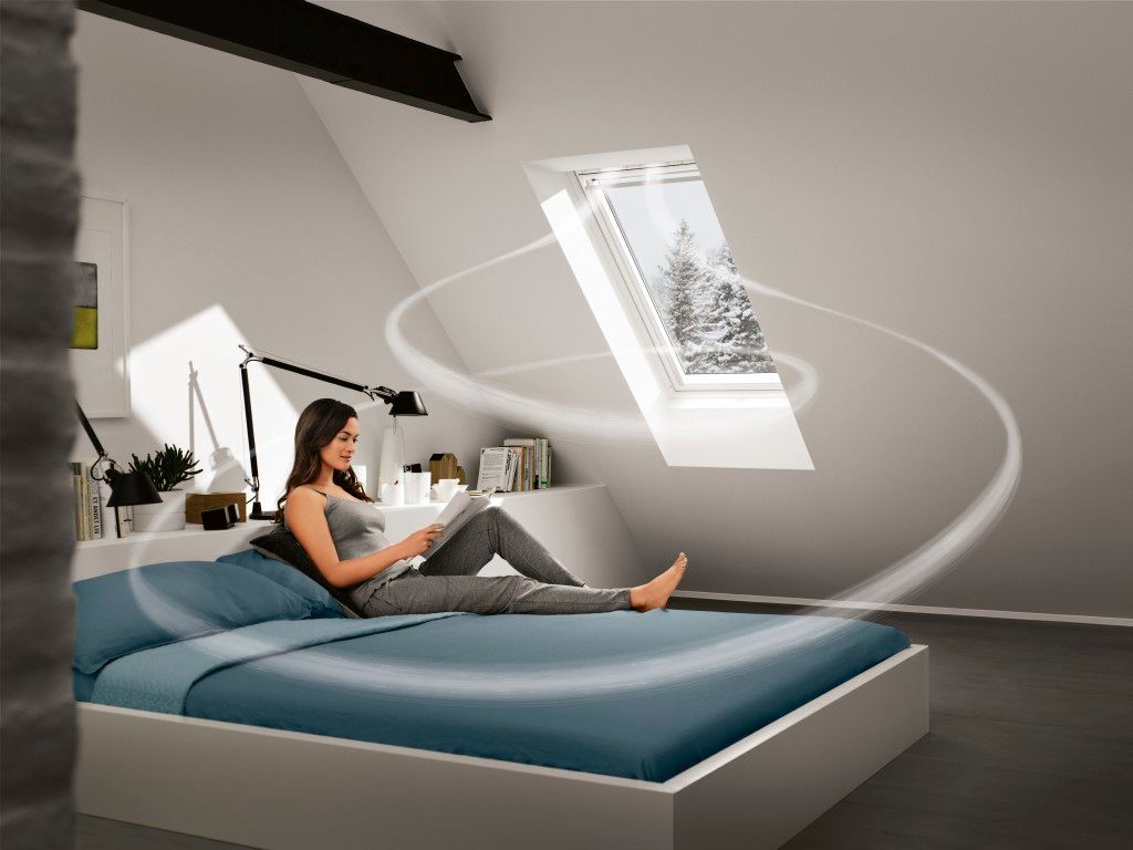 Eine Frau liegt entspannt auf dem Bett und liest ein Buch, während die Sonne durch ein Dachfenster in das Zimmer strömt und eine gemütliche Atmosphäre schafft.
